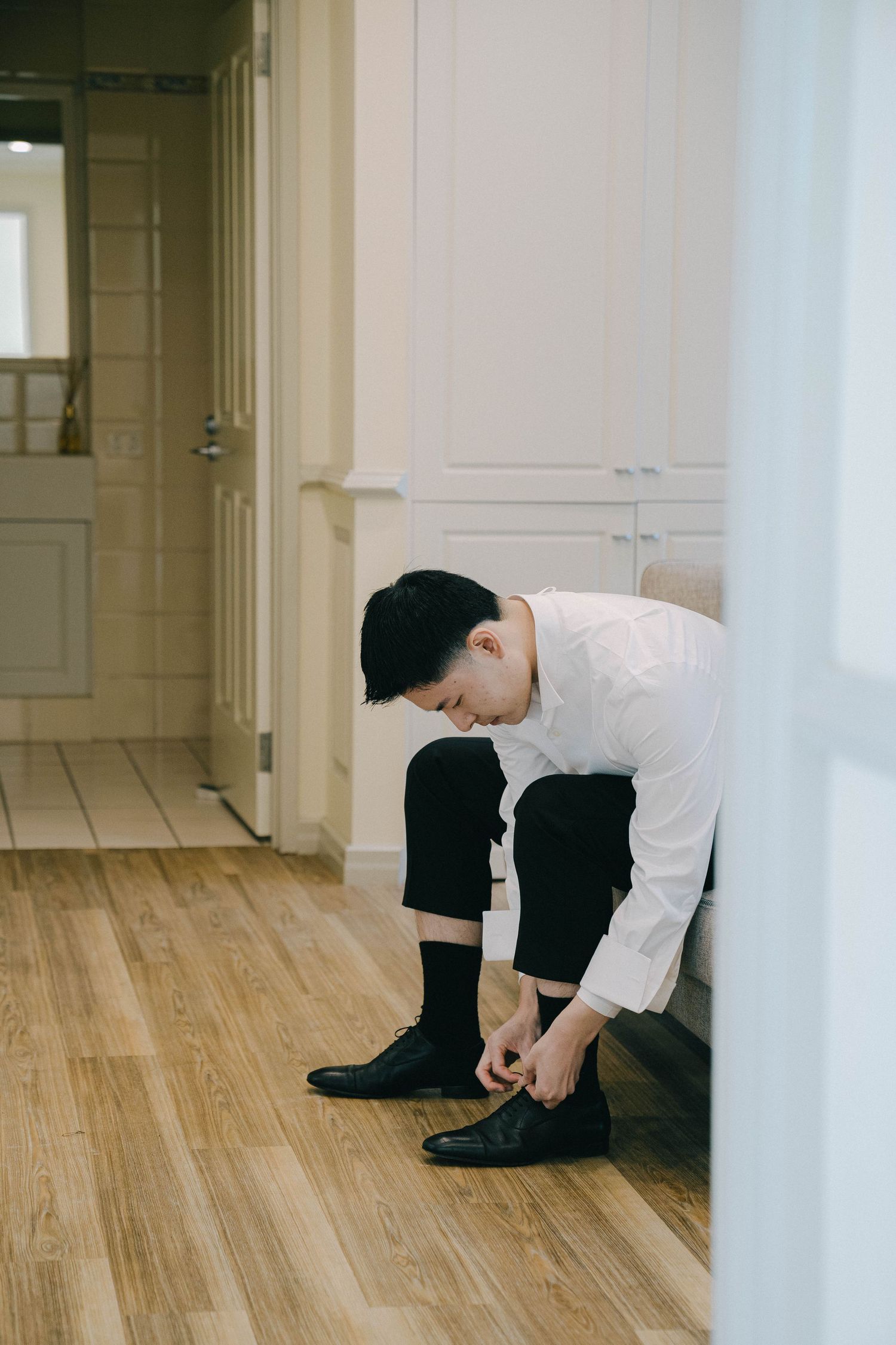 Person in white shirt and black pants sits on wooden floor in minimalist hallway with white walls.