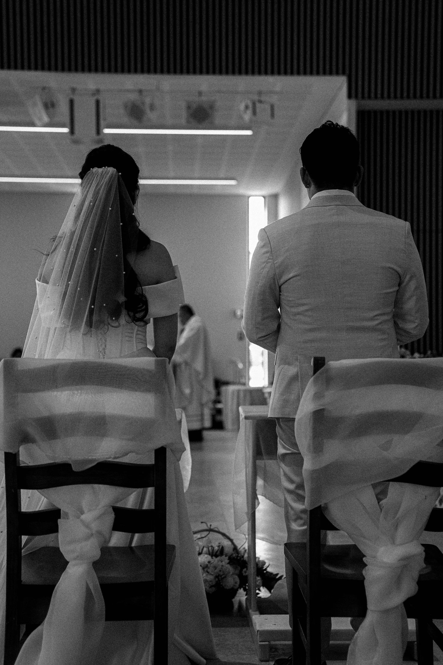 A black and white photo of a bride and groom sitting in decorated chairs during their wedding ceremony.