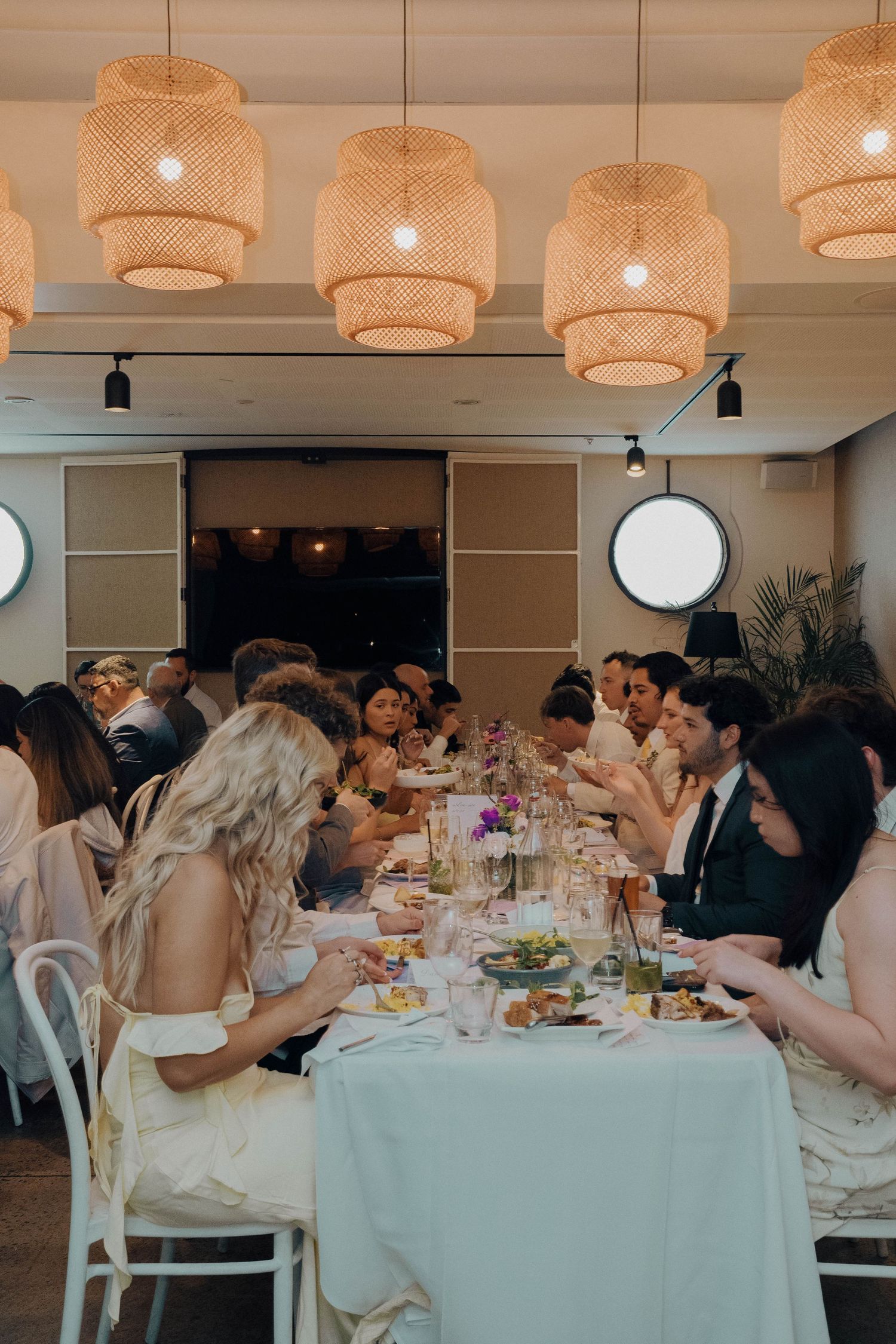 Wedding guests seated at long tables with wicker pendant lights overhead enjoy a reception dinner in an elegant venue.