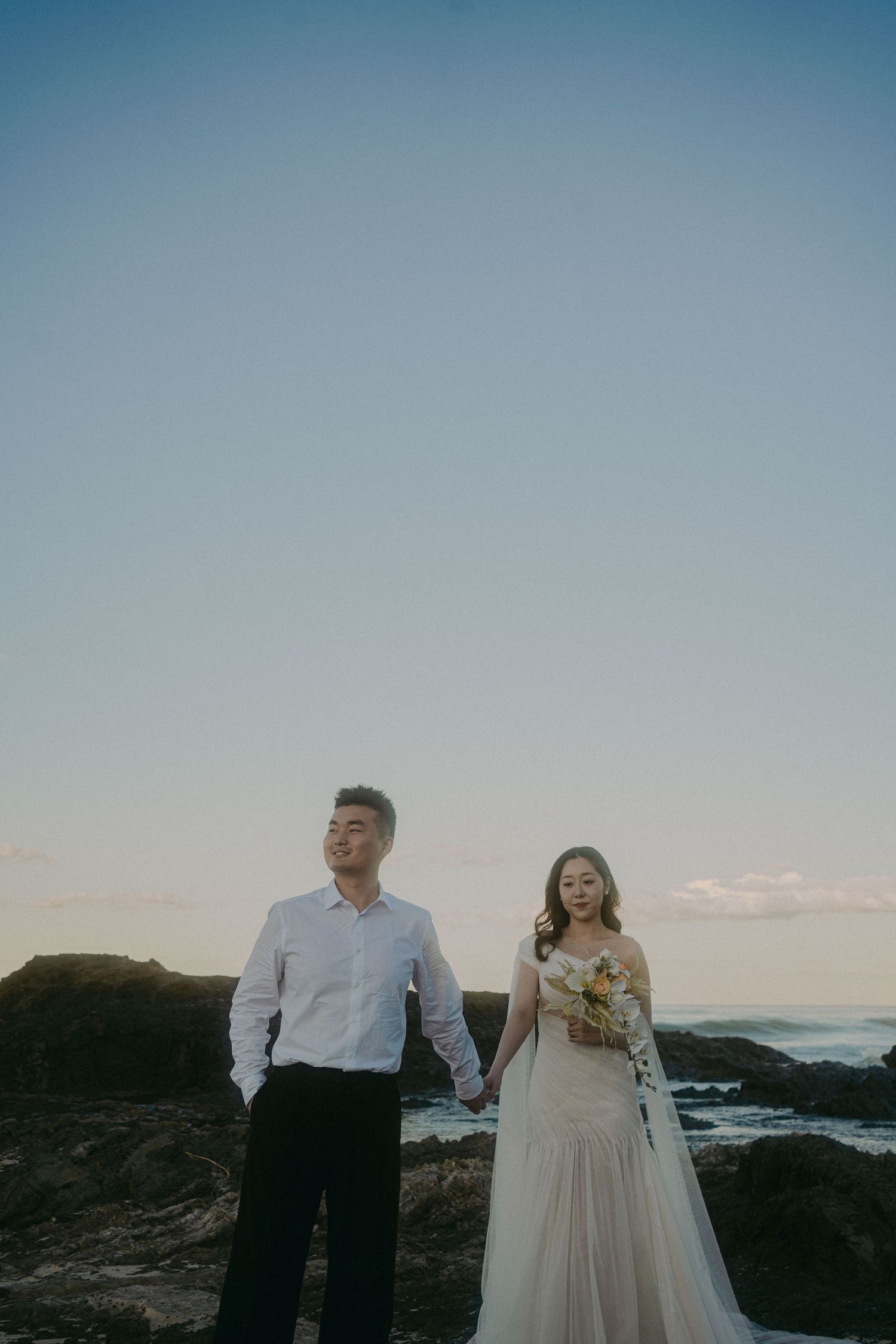 A dreamy oceanside wedding photo captured at sunset with newlyweds against dramatic coastal cliffs and crashing waves.