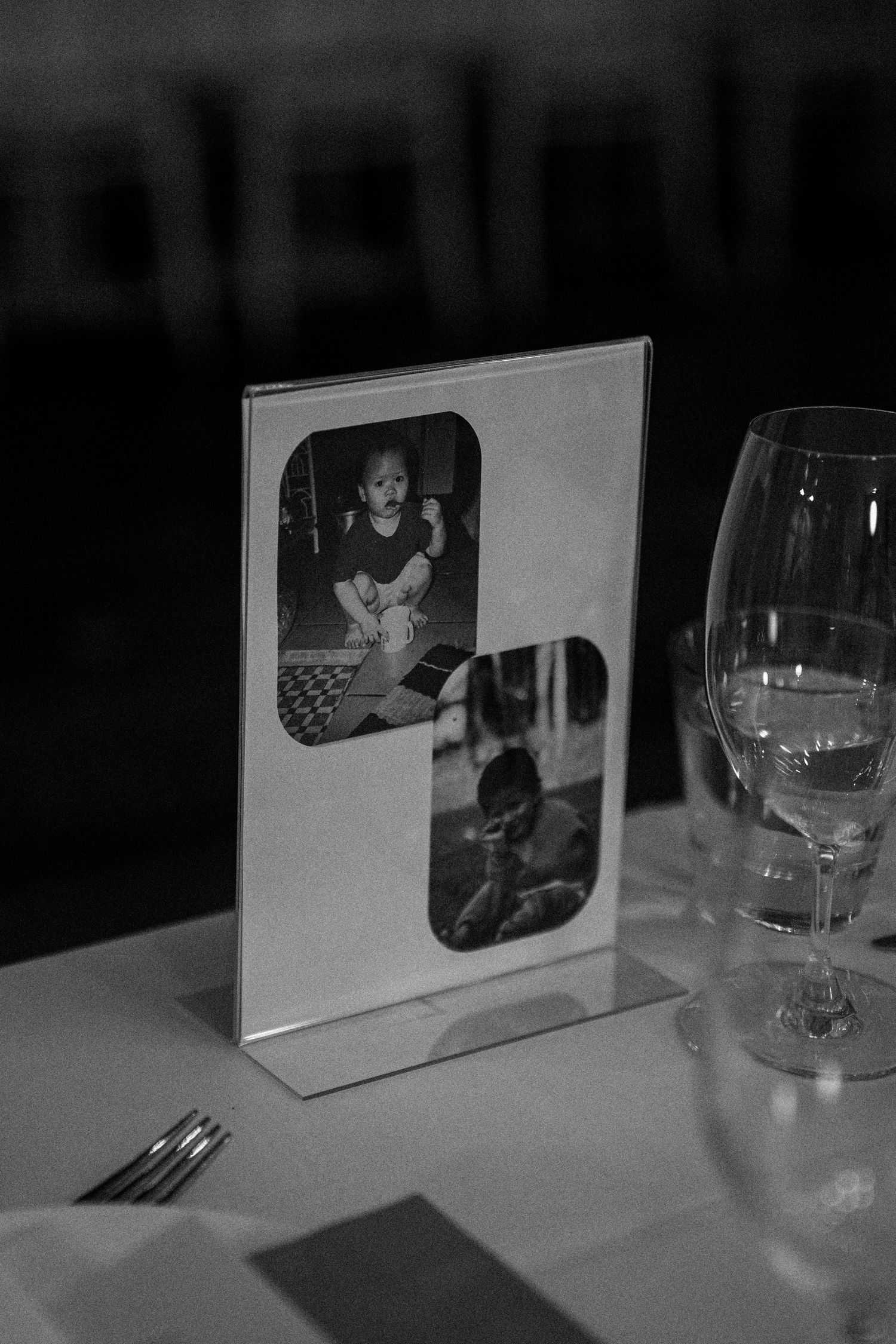 A black and white photo frame sits on a table next to wine glasses and silverware in an elegant dining setting.