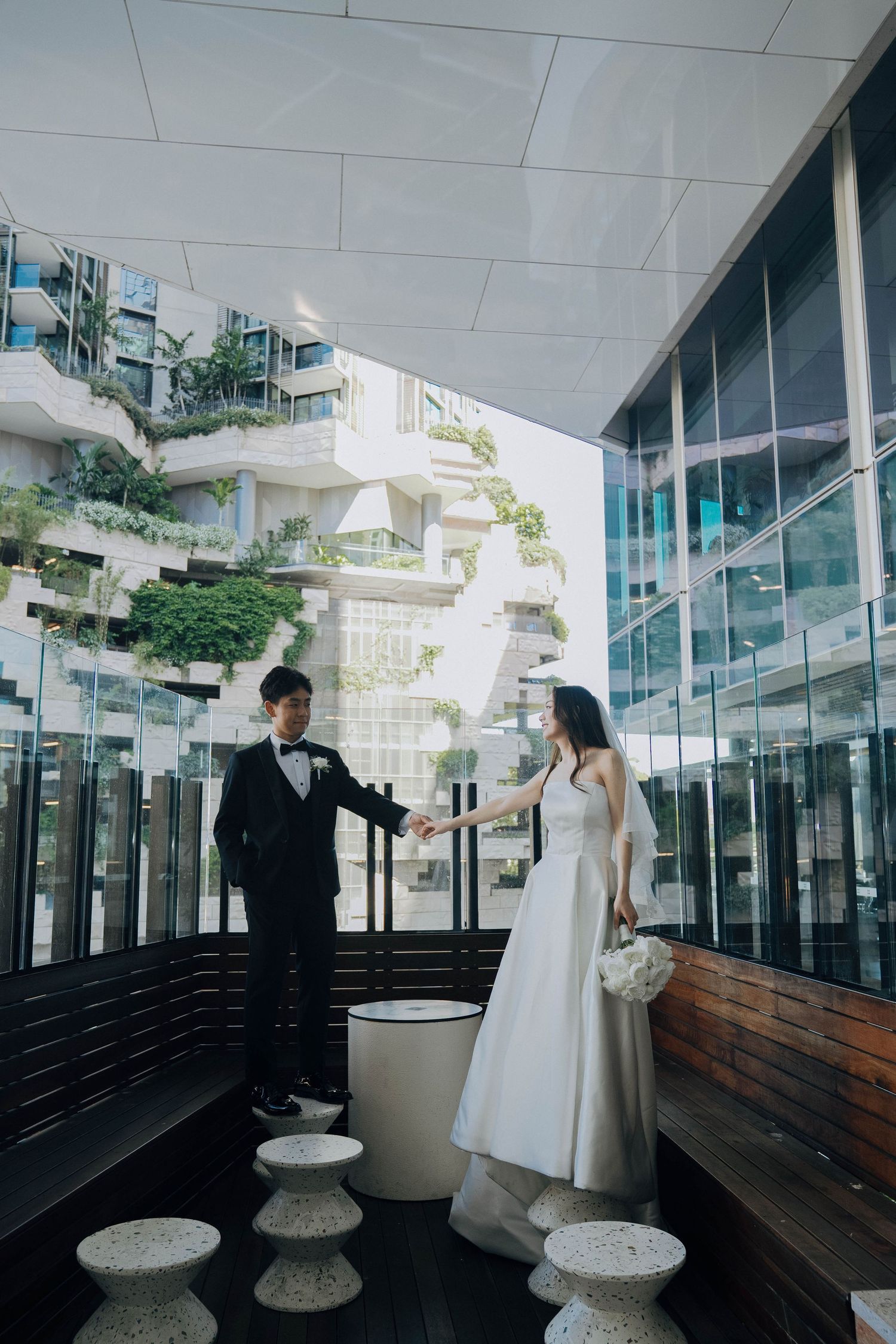 A couple in elegant attire poses together on a modern balcony with glass walls and wooden flooring.
