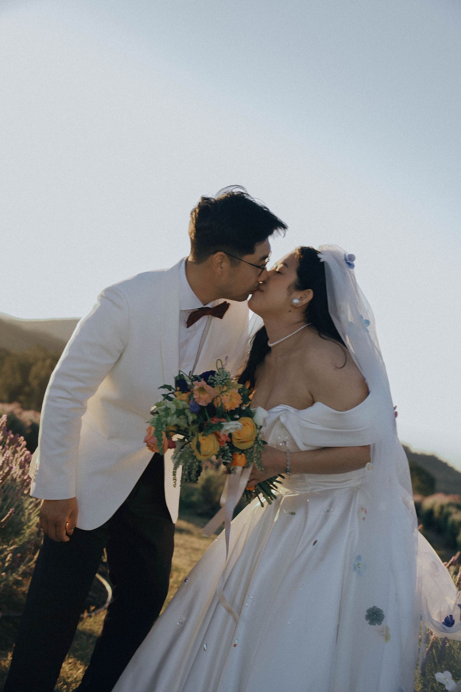Newlywed couple sharing a romantic kiss outdoors with bride holding autumn bouquet against mountainous backdrop.