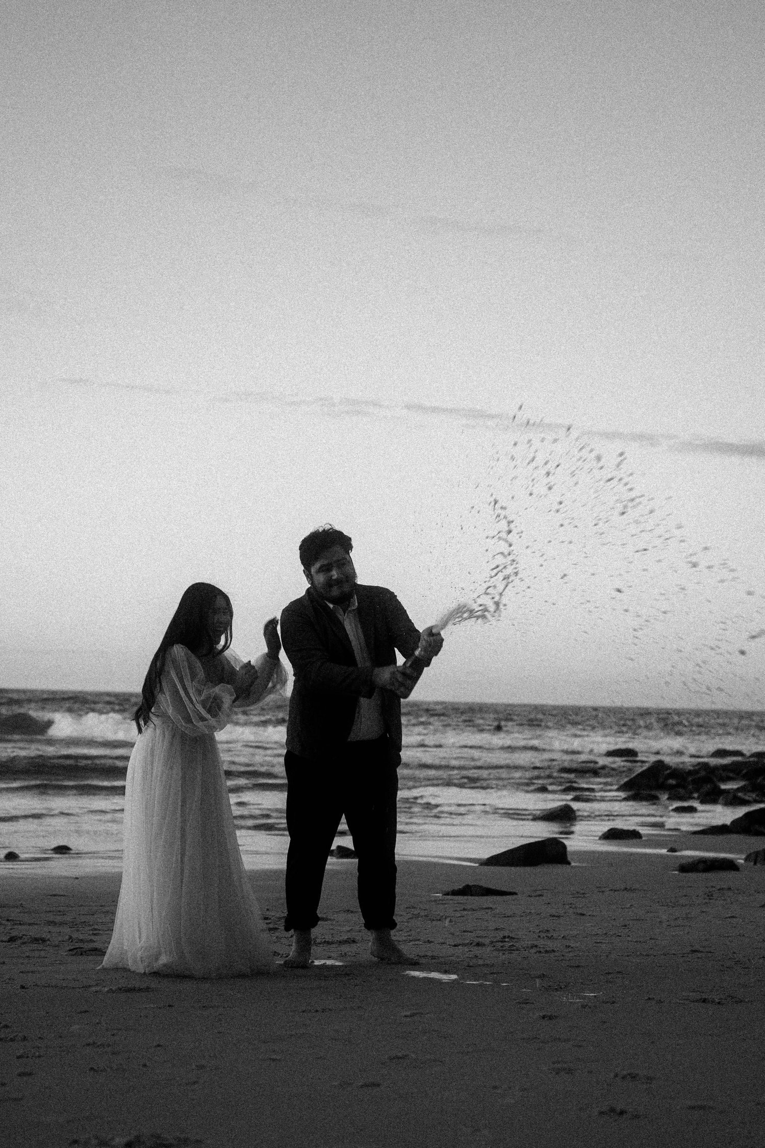 A couple in wedding attire stands on a beach while a flock of birds soars overhead in black and white photography.