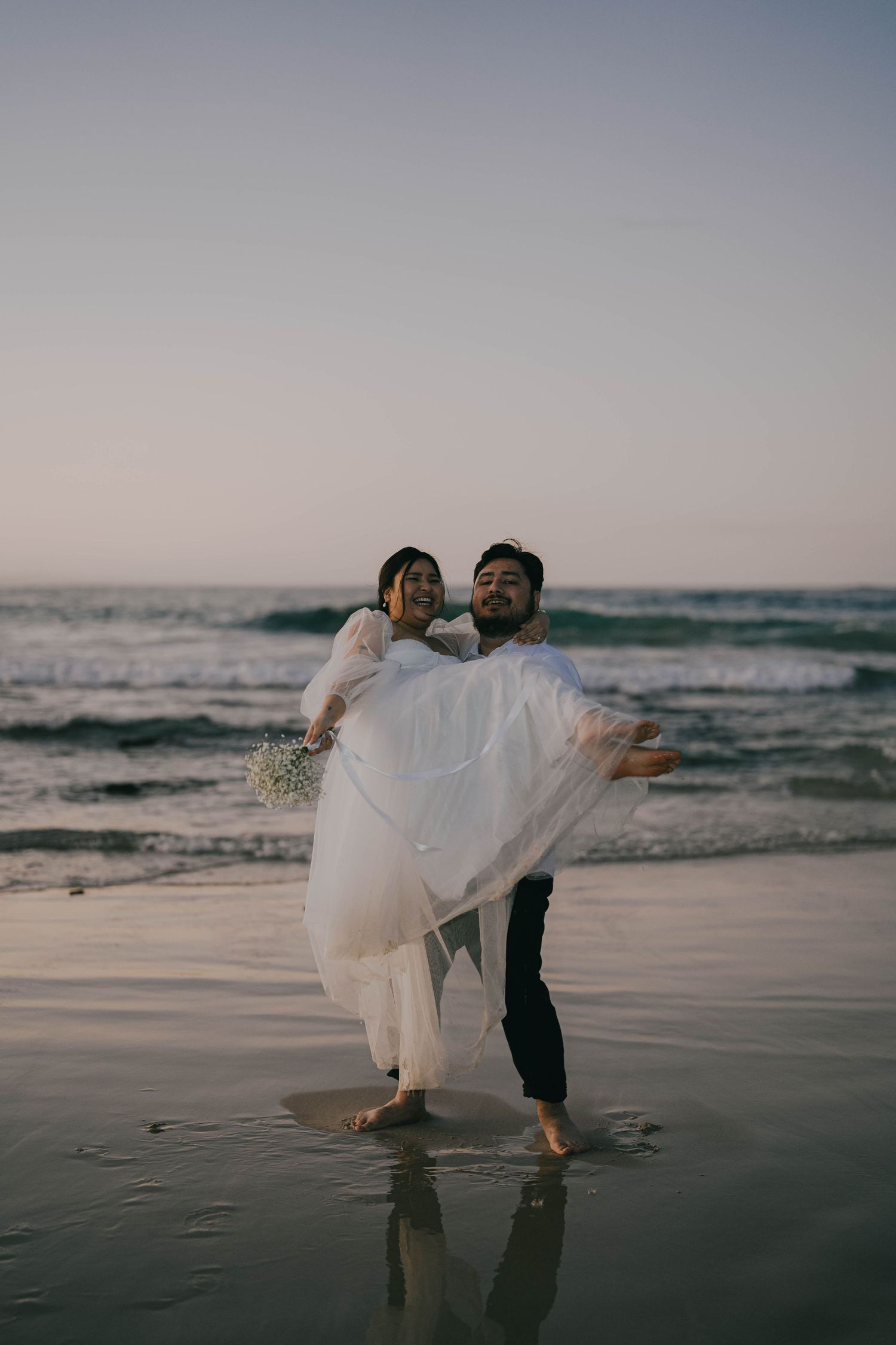 A couple in wedding attire dances and embraces on a serene beach at sunset with ocean waves in the background.