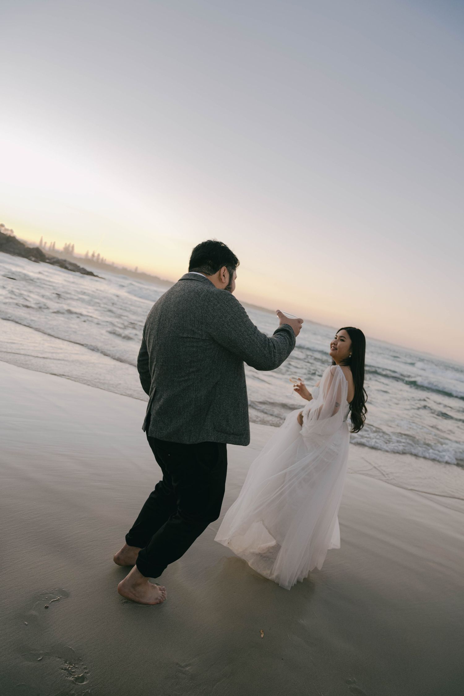 A couple in formal attire poses for romantic photos at sunset on a beach with waves in the background.