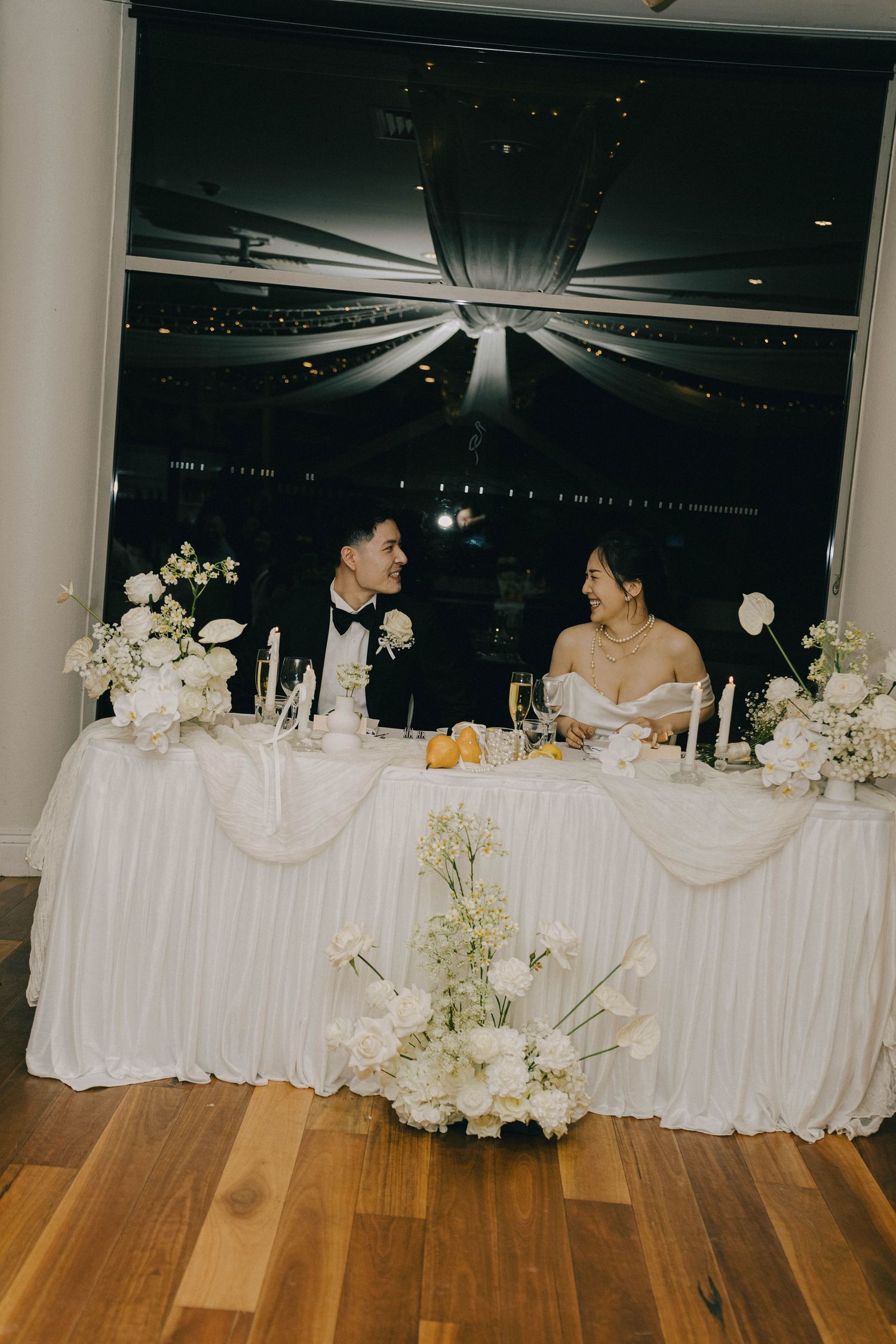 A wedding reception head table decorated in white linens with floral arrangements sits atop a wooden dance floor.