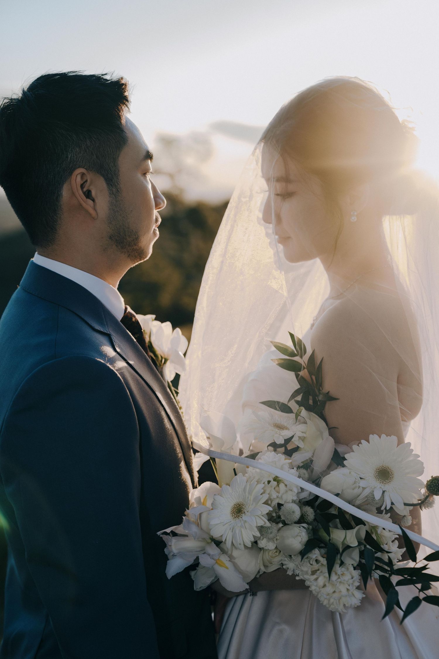 A romantic wedding portrait of a couple with white flowers and sunlight through a veil in an outdoor setting.