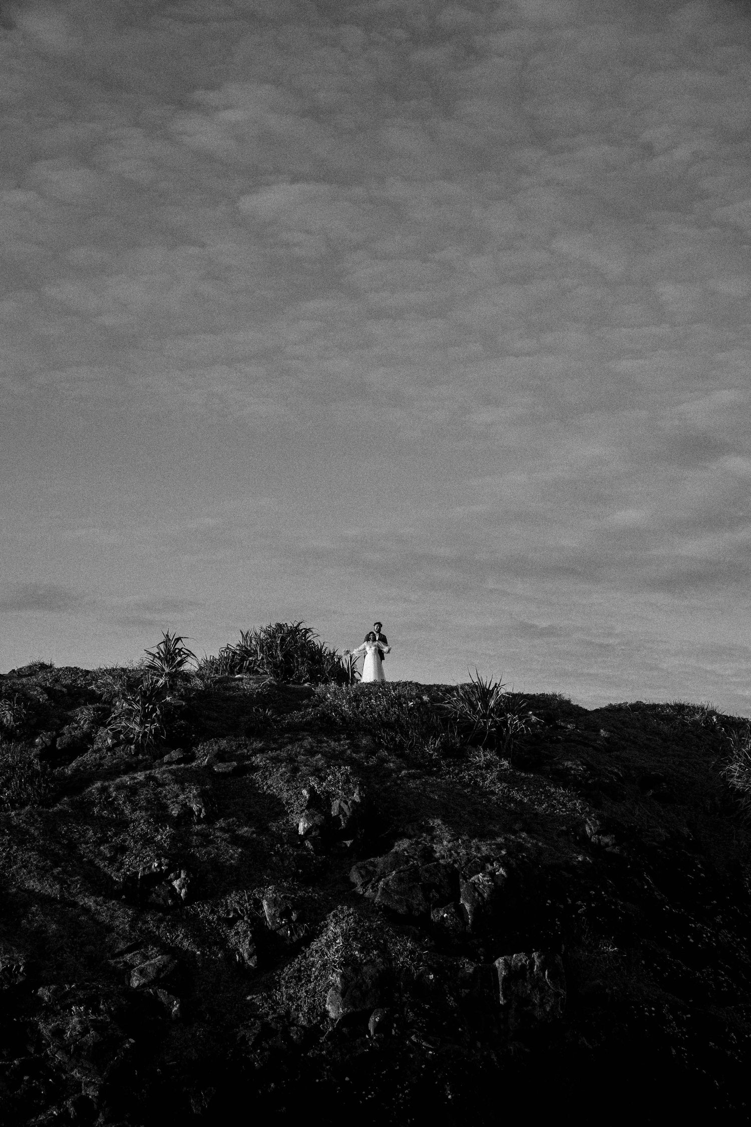 A lighthouse stands silhouetted against a cloudy sky on a dark hilltop surrounded by dense vegetation.
