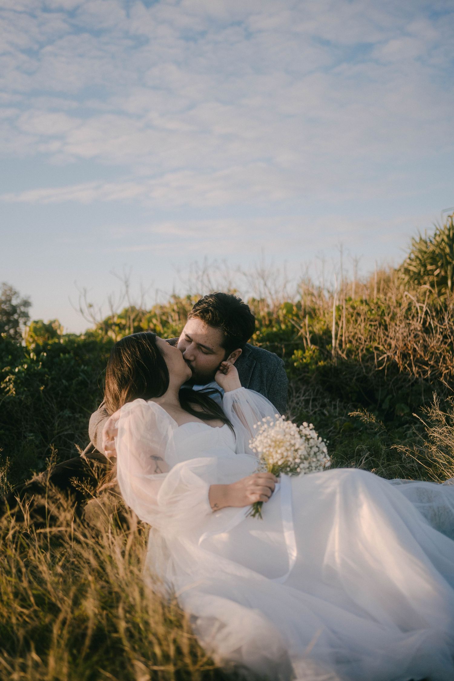 A couple in wedding attire reclines in golden grass at sunset while sharing a romantic moment.