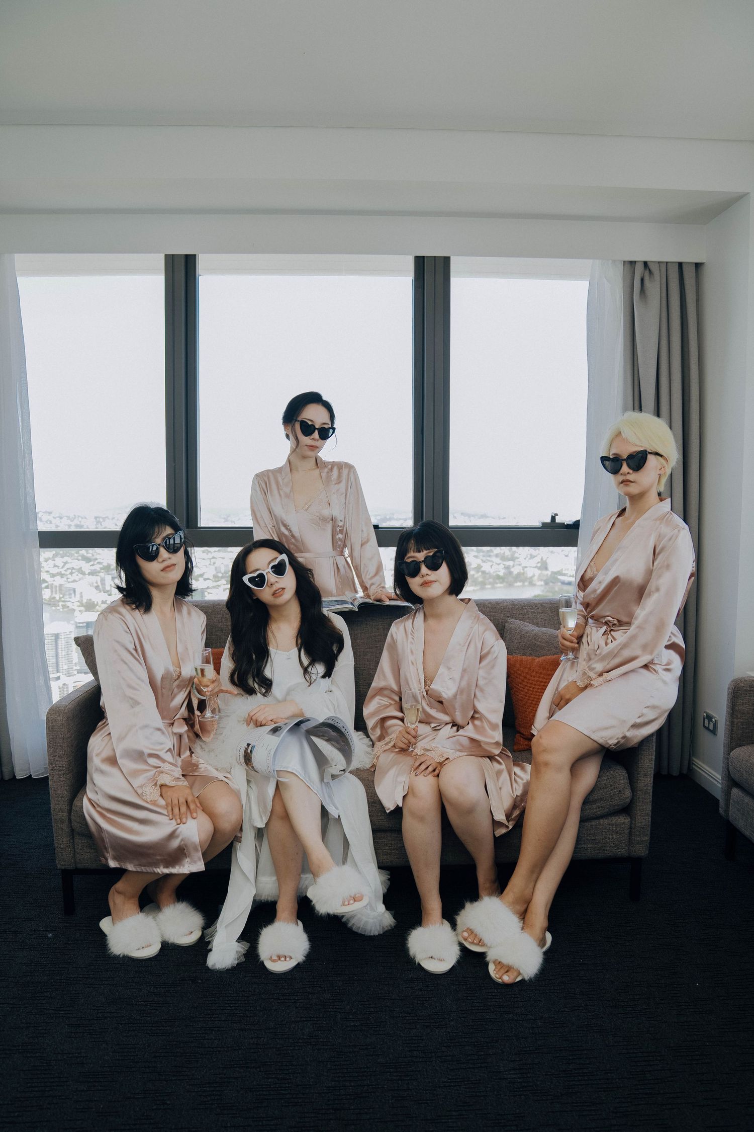 Four people in matching pink satin robes and sunglasses sit together on a bench in a modern room with windows.