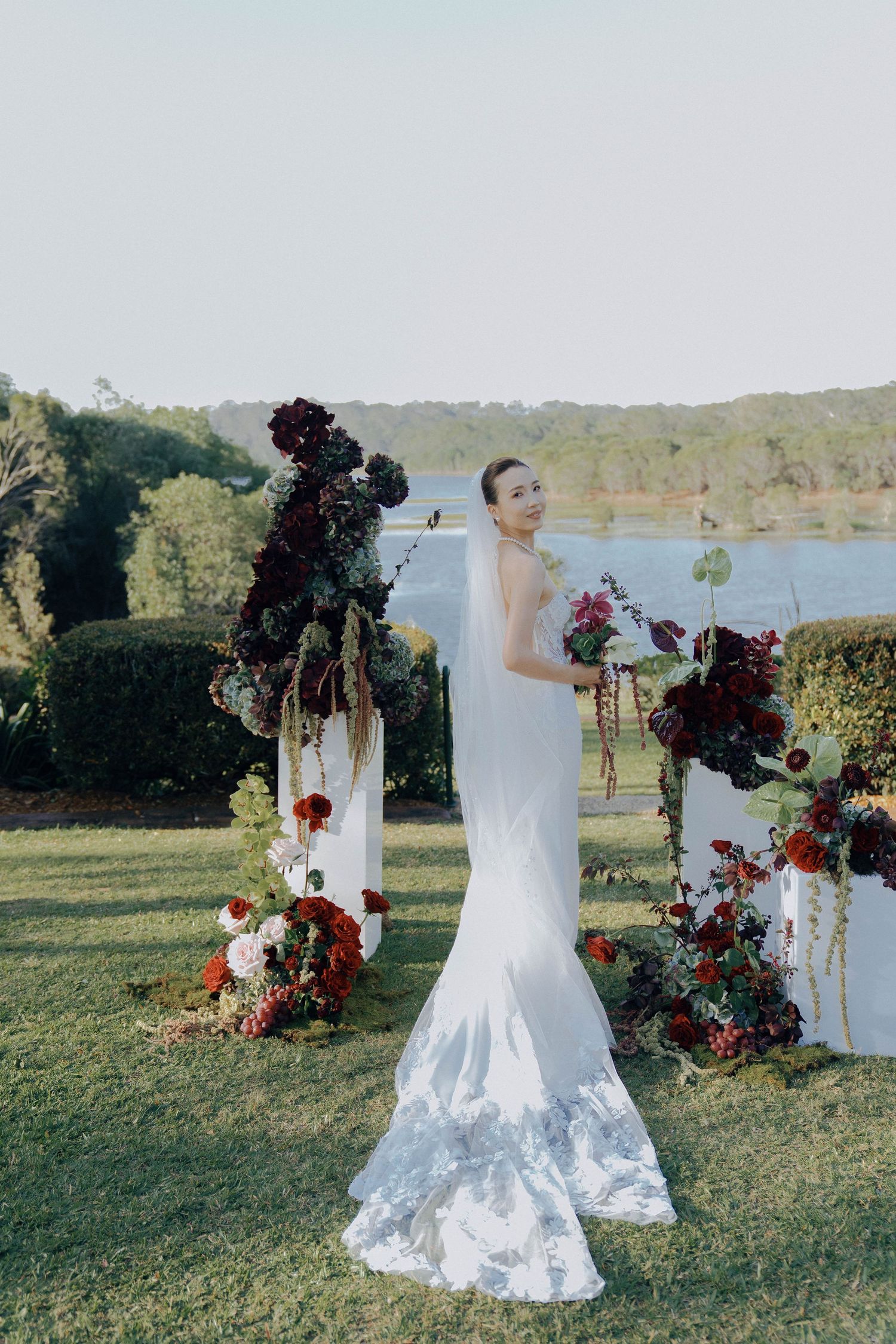 A white wedding dress displayed outdoors with burgundy floral arrangements against a lakeside backdrop.