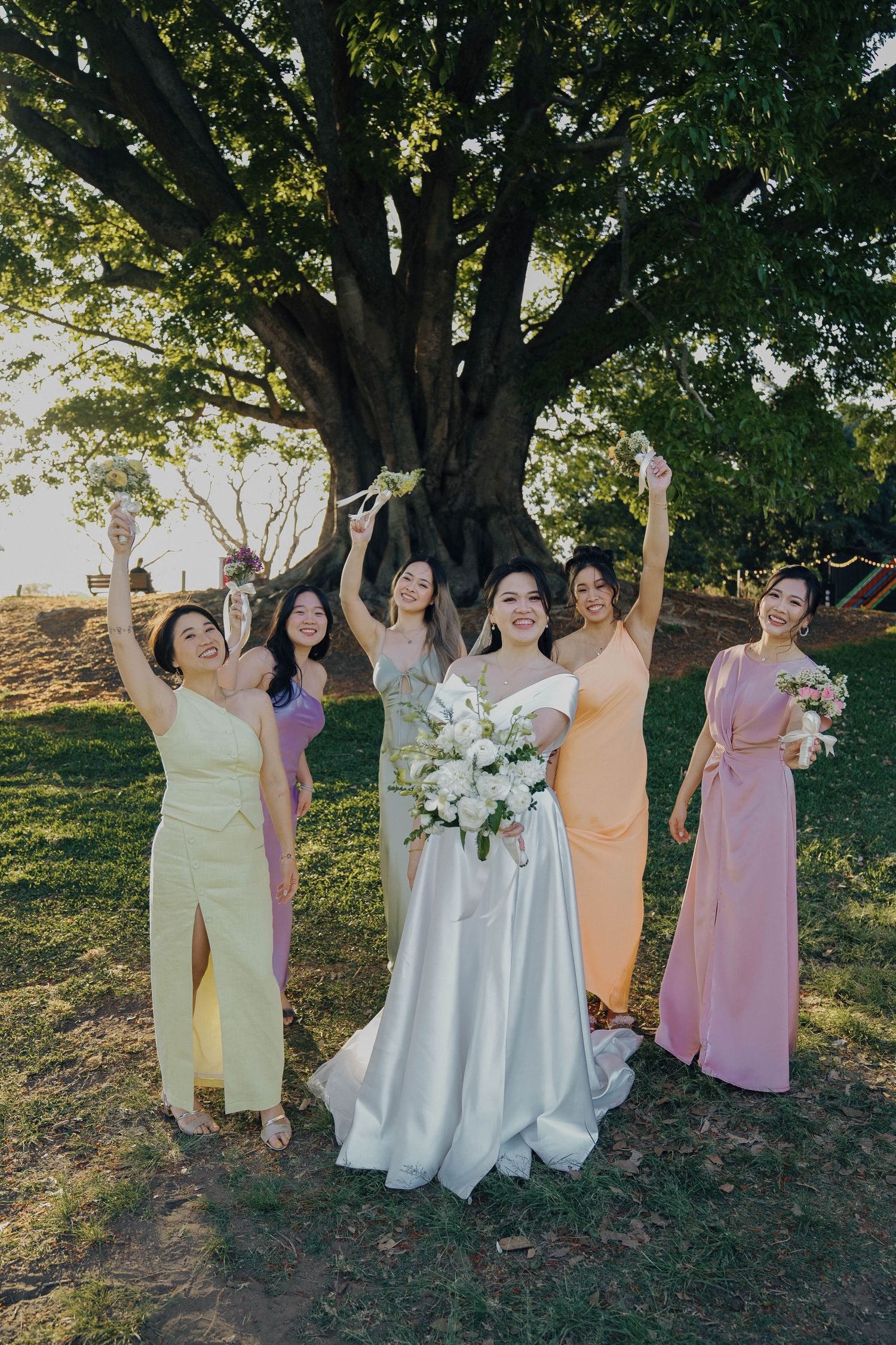 Group in pastel dresses pose joyfully together under trees during sunset at outdoor wedding celebration.