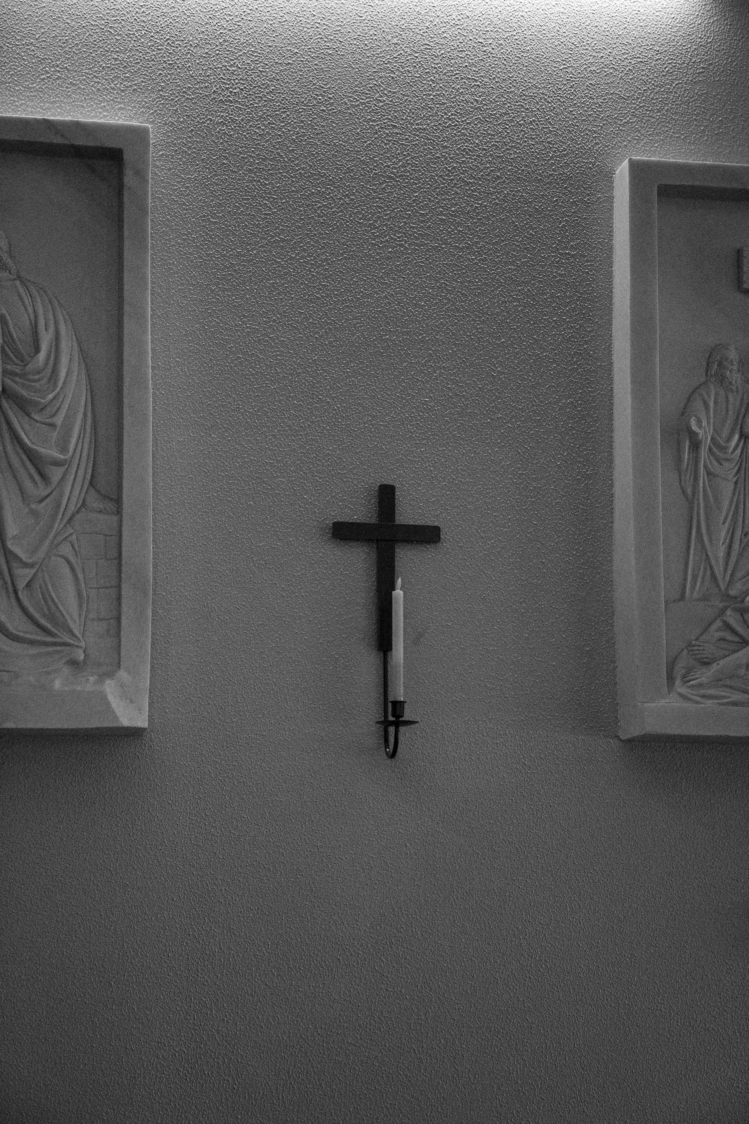 Black and white photographs show a simple cross mounted on a minimalist church wall with religious artwork panels.