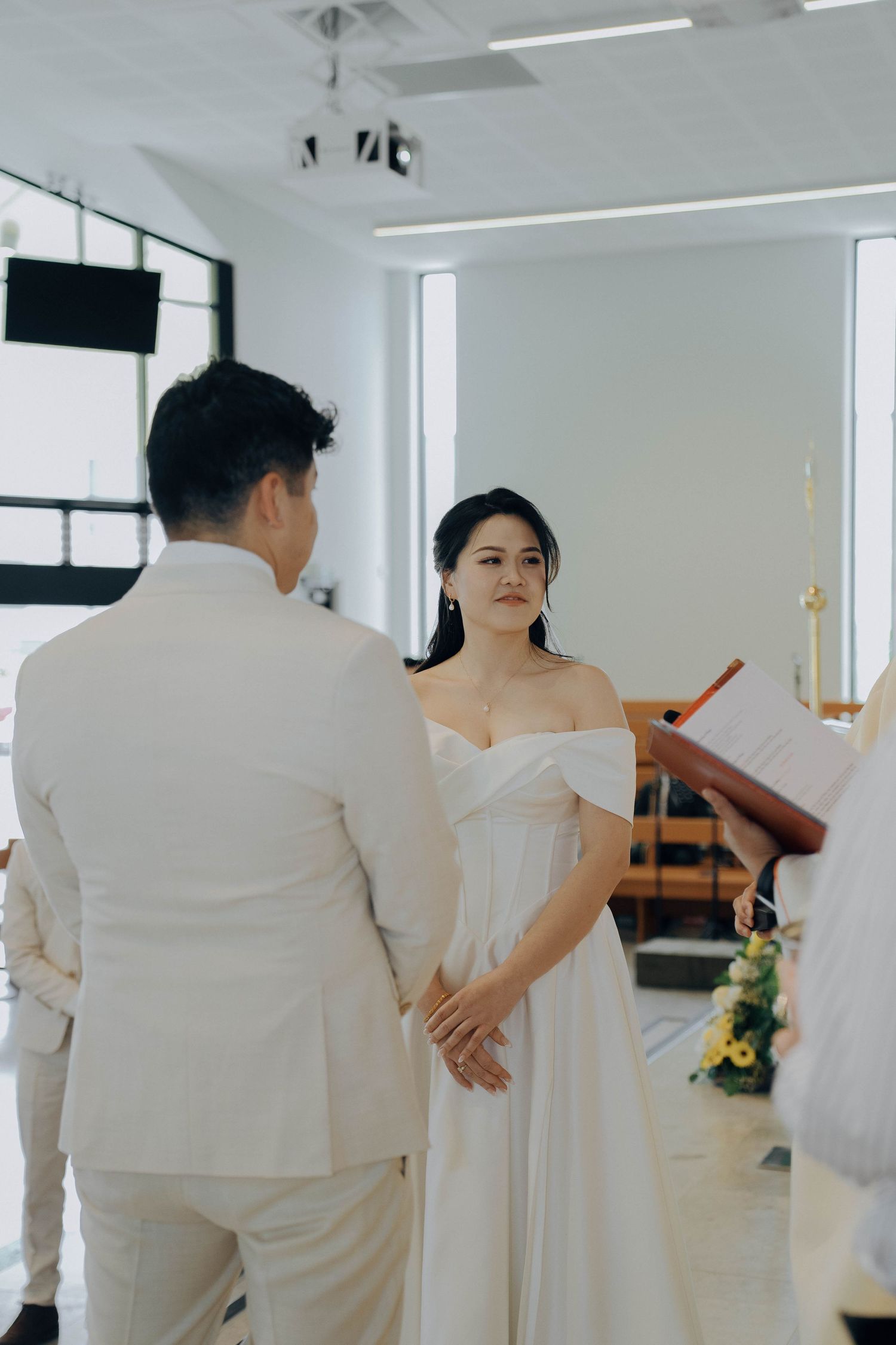 A wedding ceremony takes place in a bright white modern chapel with minimal decor and tall windows.