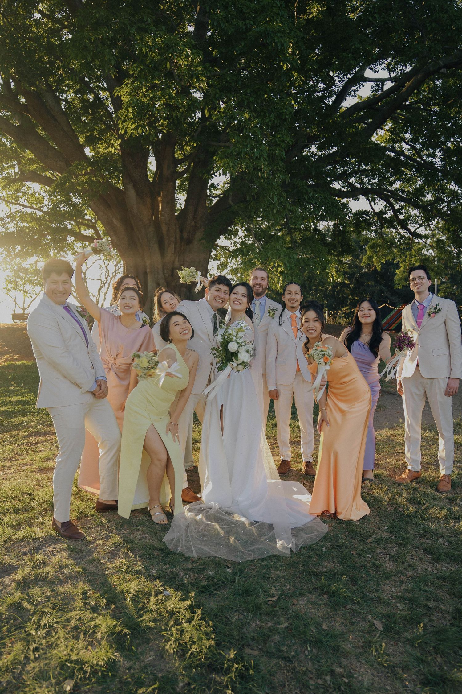 Wedding party poses together outdoors at sunset under a large tree in pastel attire.