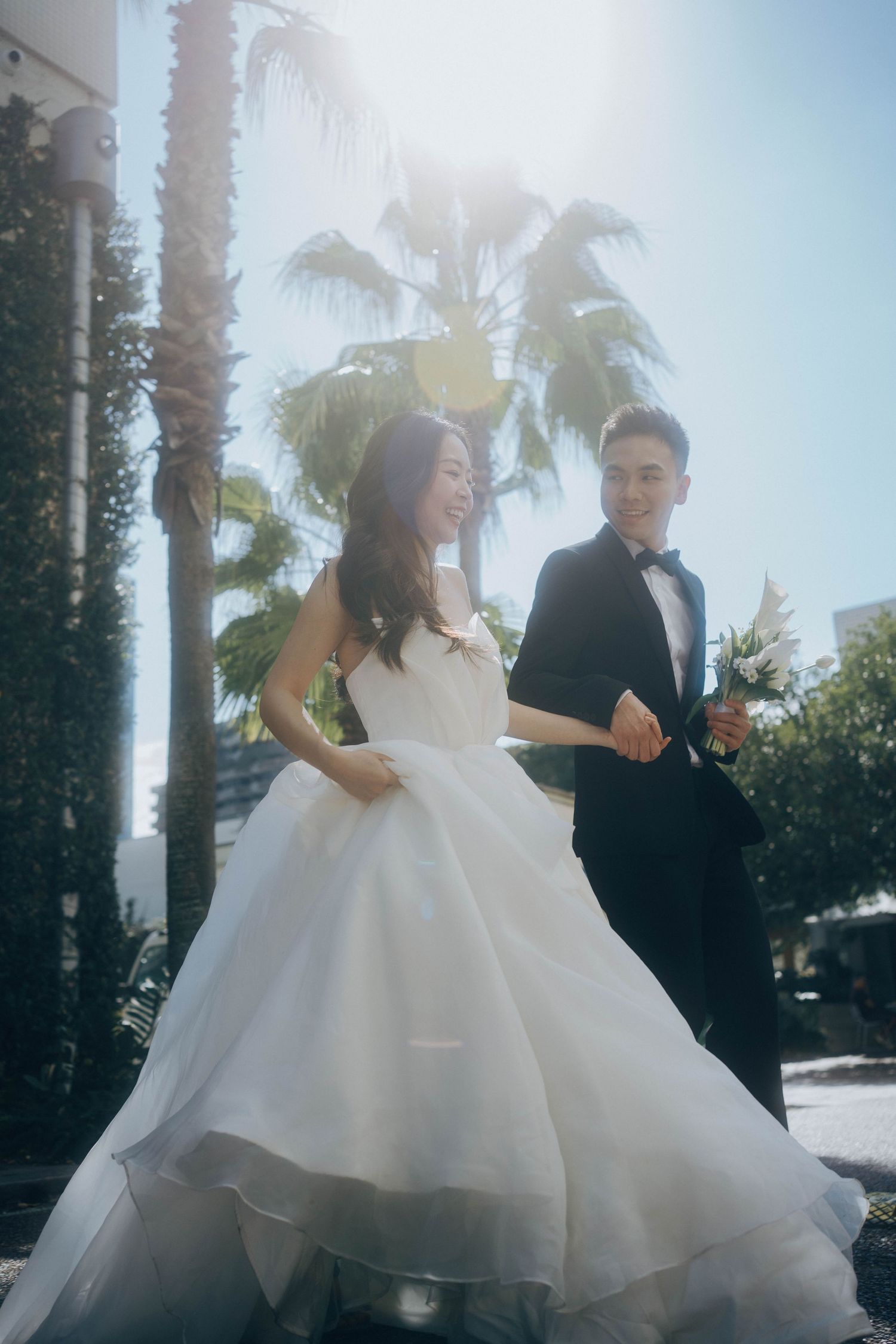 A bride in a flowing white ballgown and groom in black suit dance together among palm trees on their wedding day.