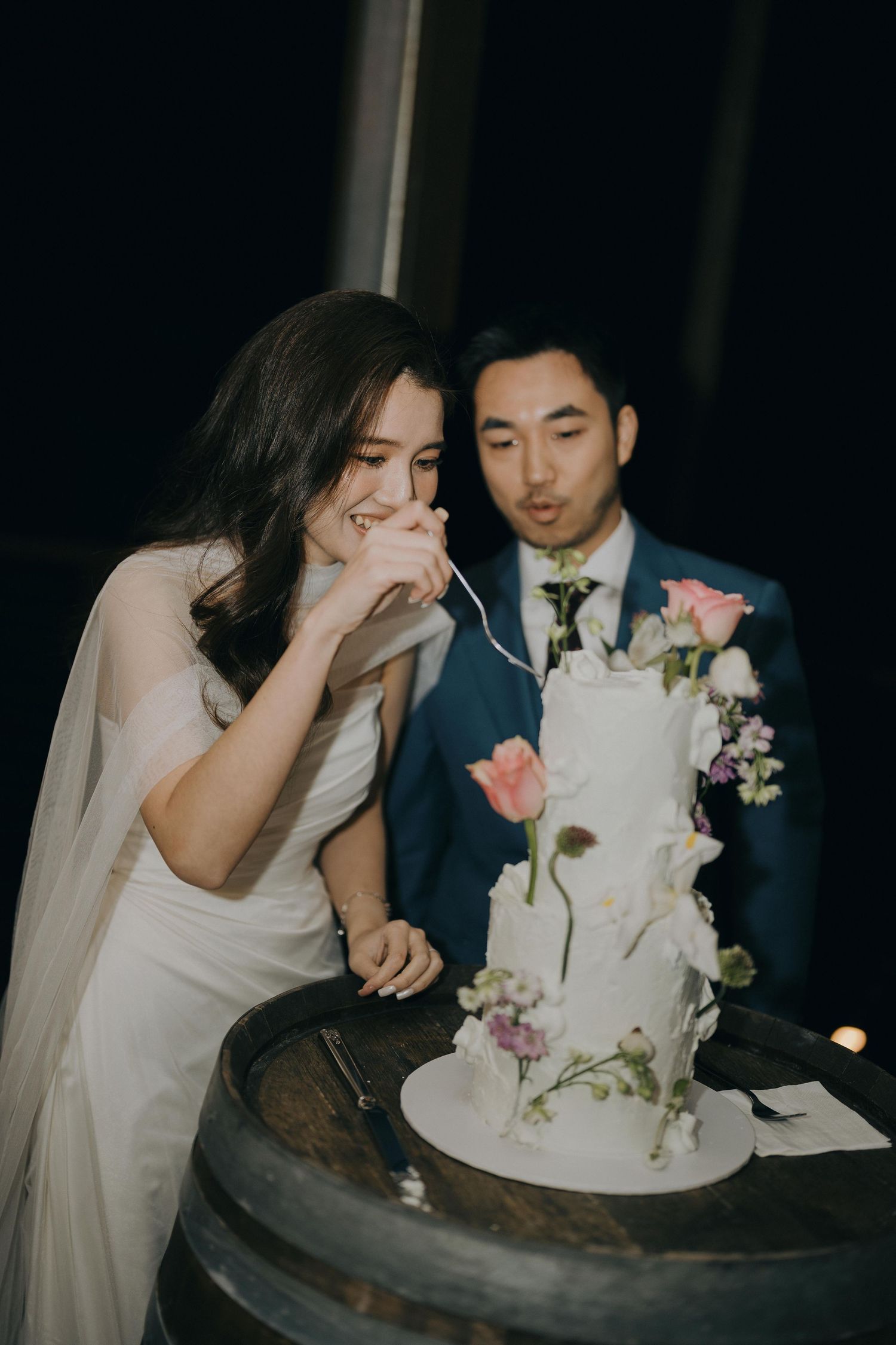Wedding couple stands together at their elegant white tiered cake decorated with pink roses and purple flowers.
