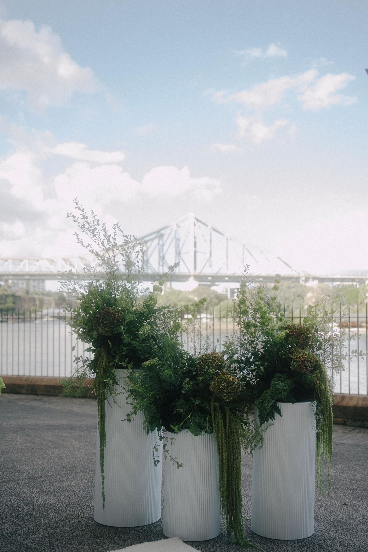 White decorative planters with cascading greenery displayed along riverfront with a large bridge visible in background.