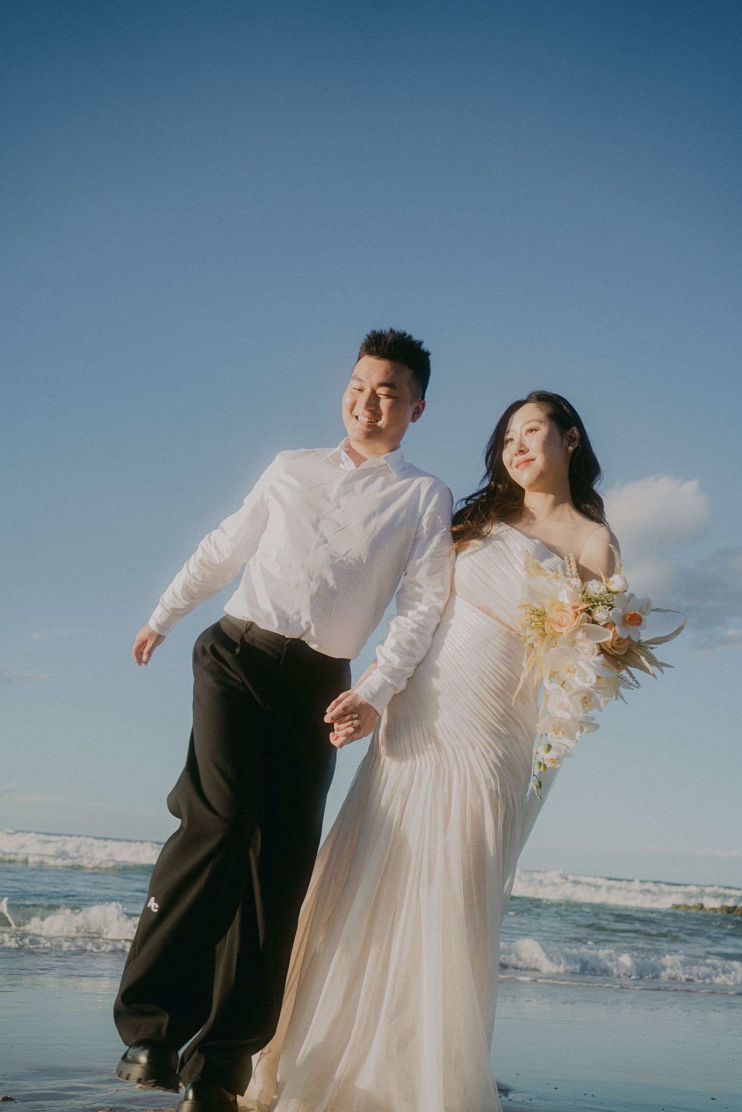 A couple in wedding attire walks joyfully along the beach with ocean waves and blue sky in the background.