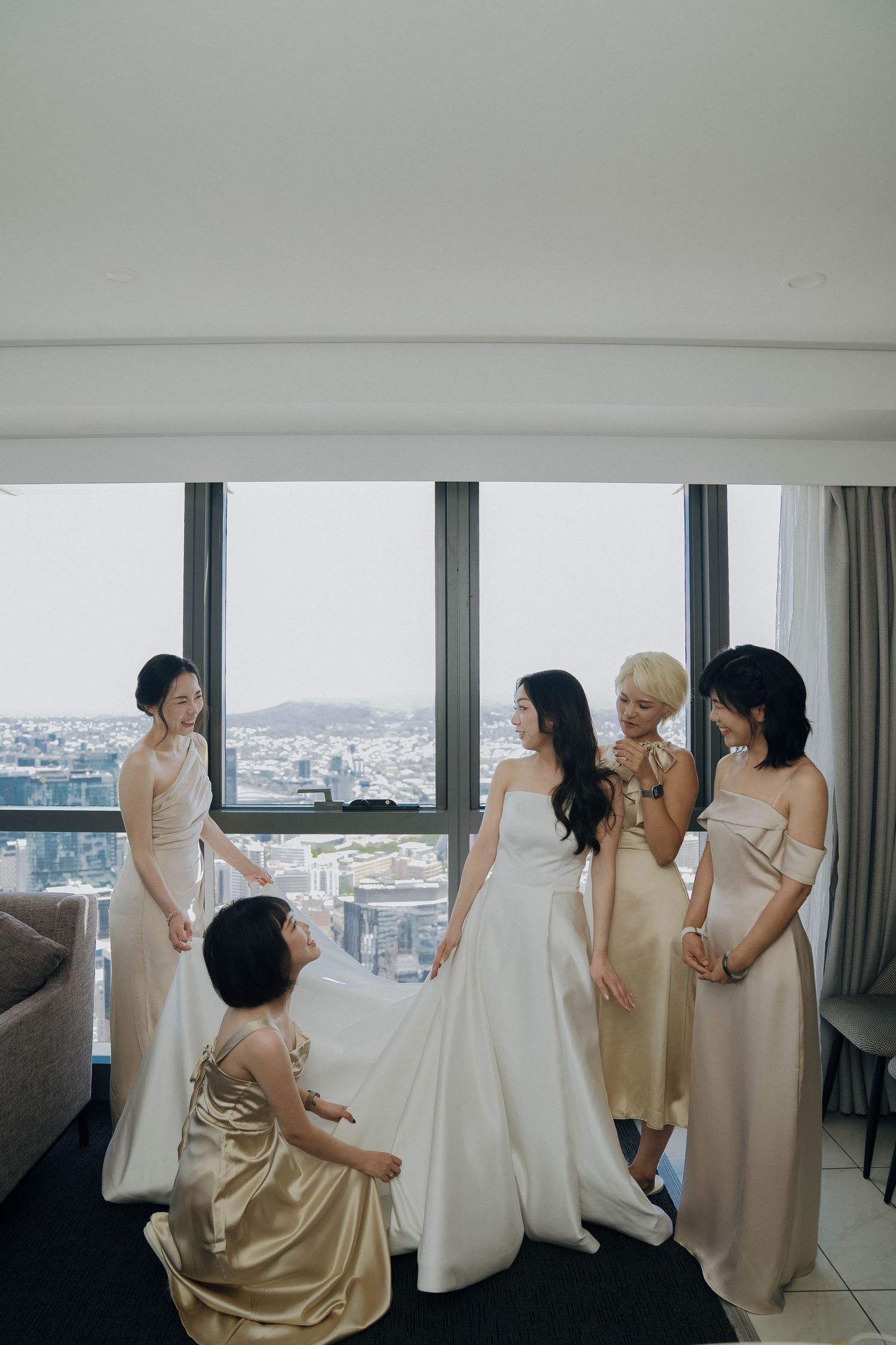 A group in cream-colored dresses gathers by large windows overlooking a city skyline.