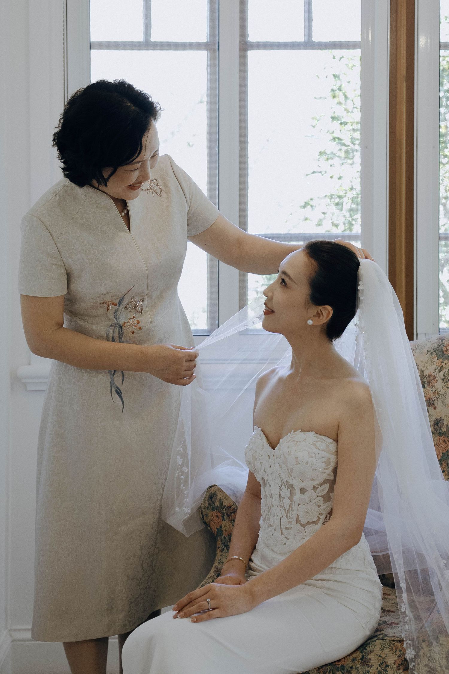 Wedding preparation scene showing bride in white gown getting ready with assistance in a bright sunlit room.