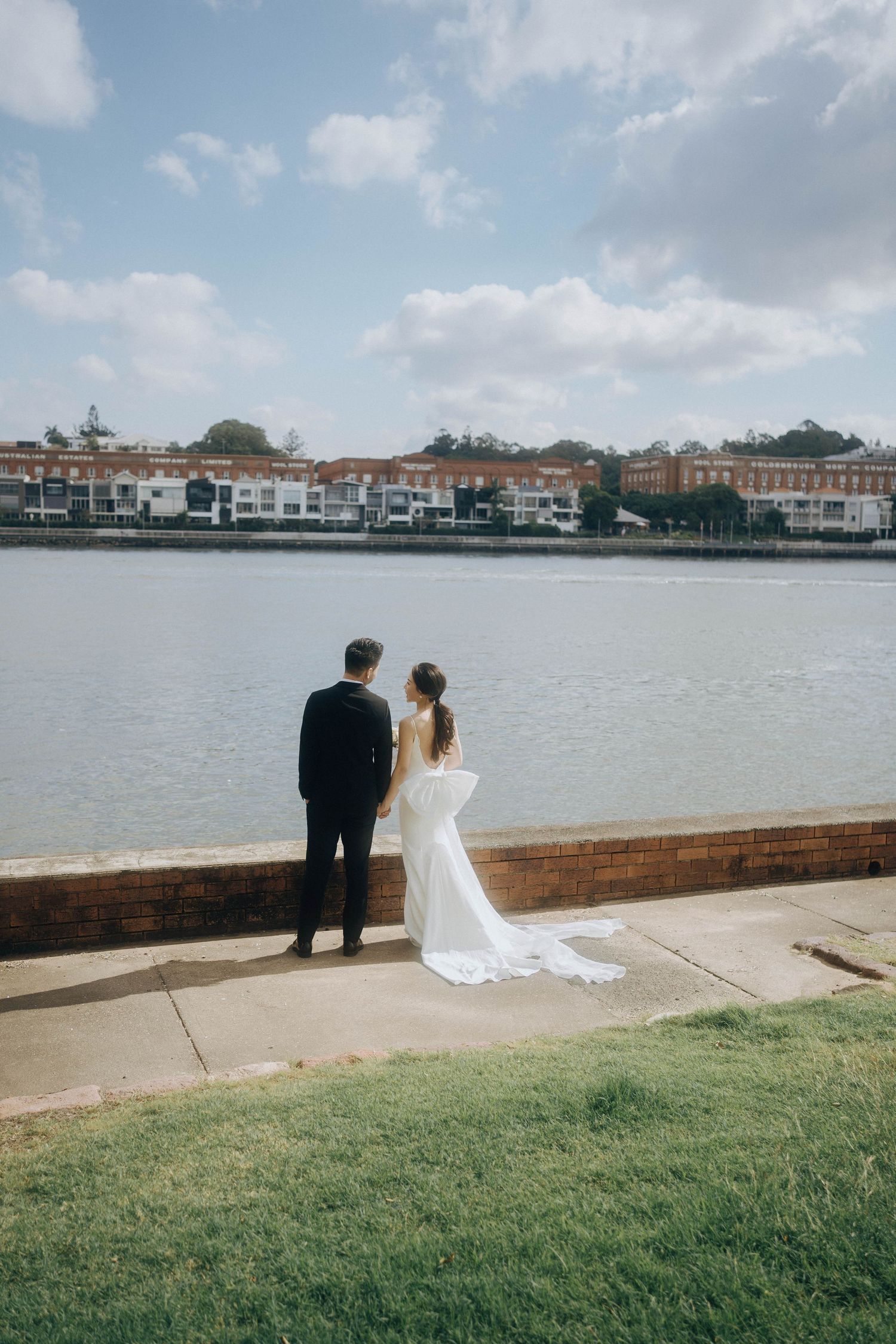 A couple in wedding attire stands at the waterfront overlooking houses and buildings across the bay on a cloudy day.