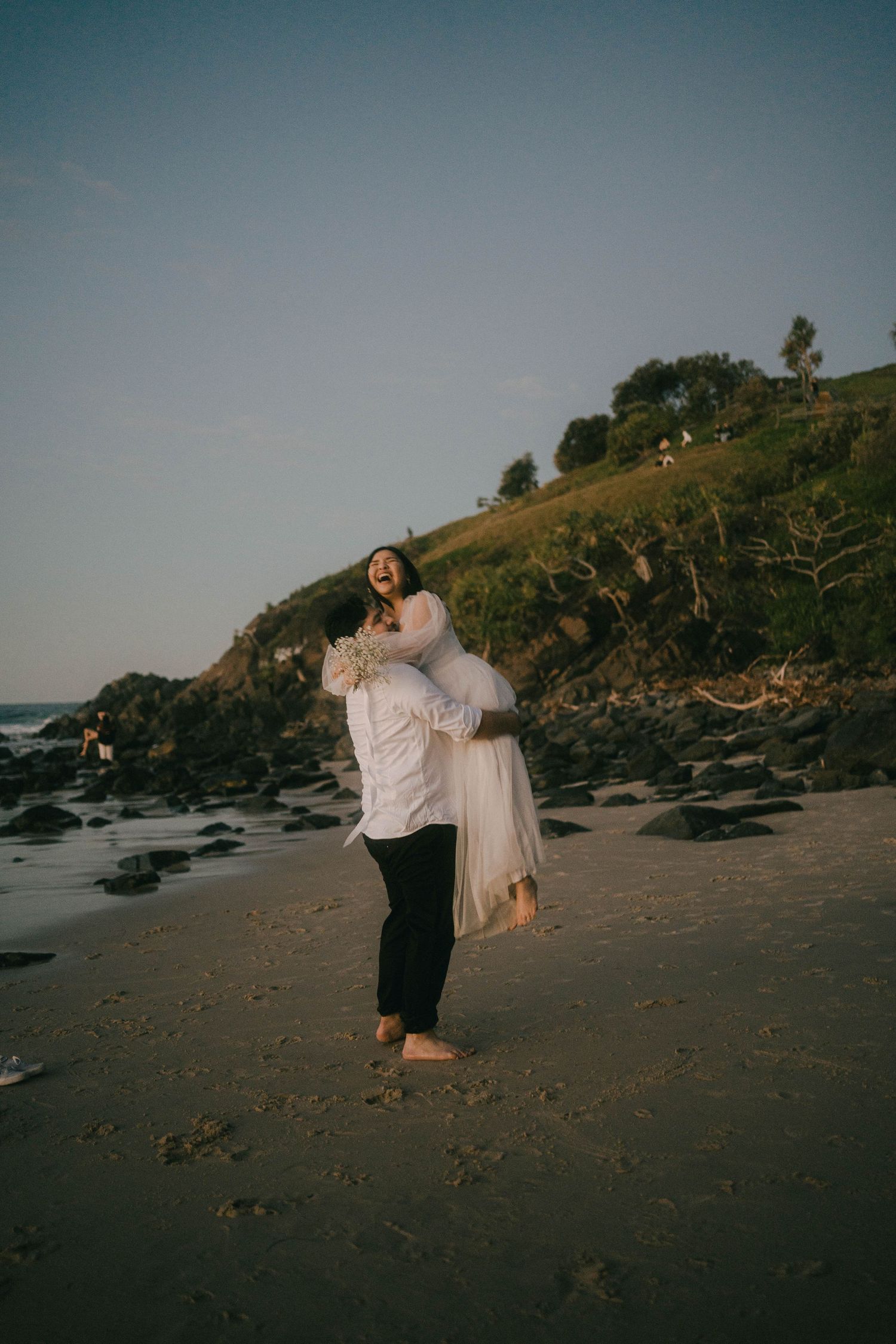 A couple dances together on a scenic rocky beach at sunset with green cliffs in the background.