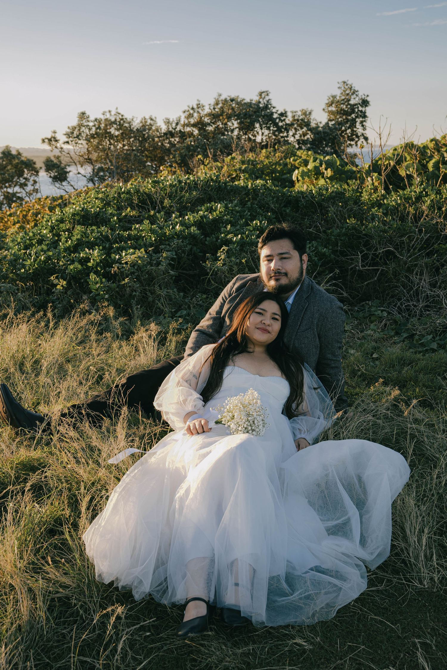 A couple in formal attire sits together on grass during golden hour sunset in a romantic outdoor setting.