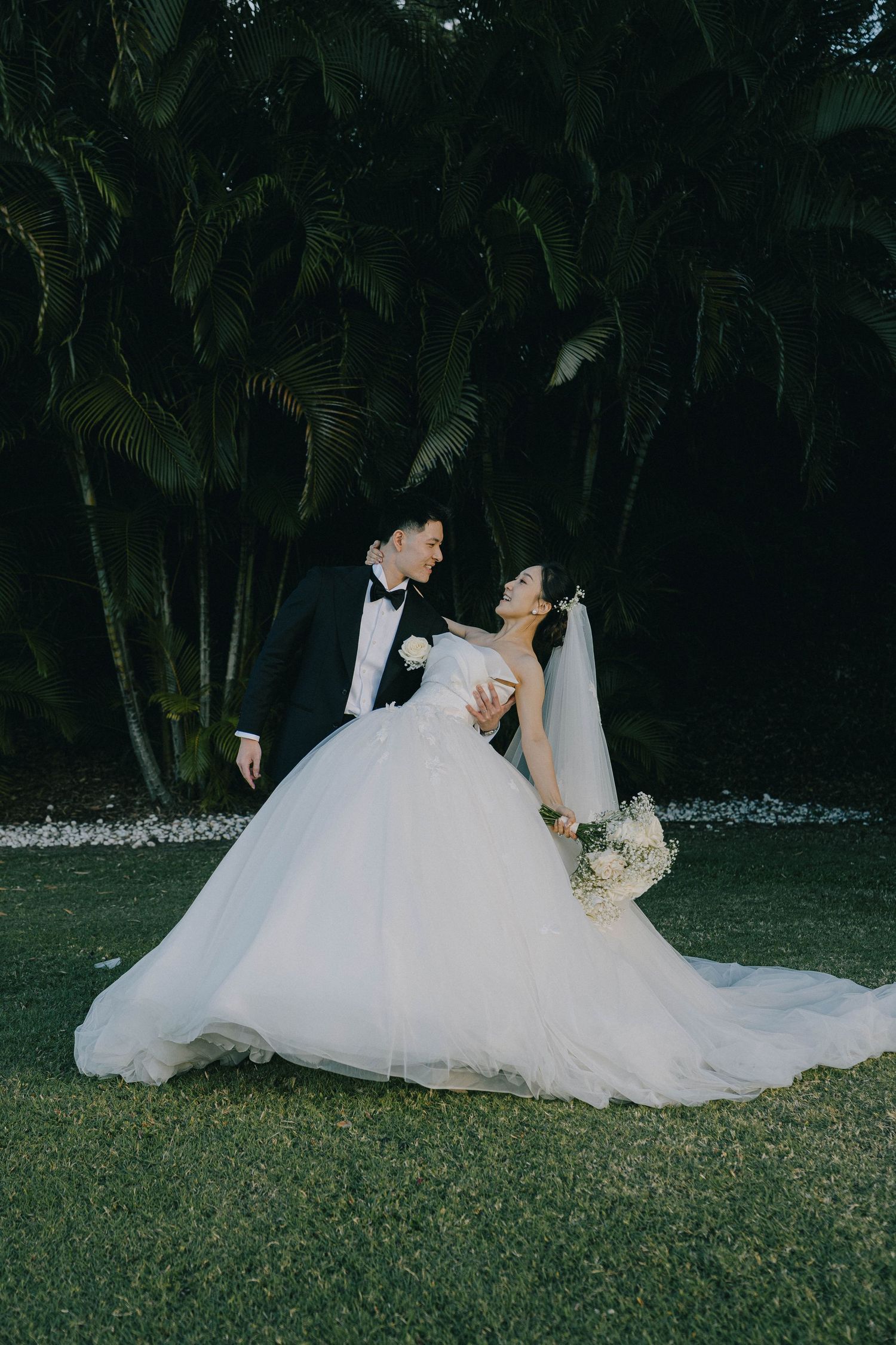 Romantic wedding couple poses on lush green lawn with tropical palm trees in evening garden setting.