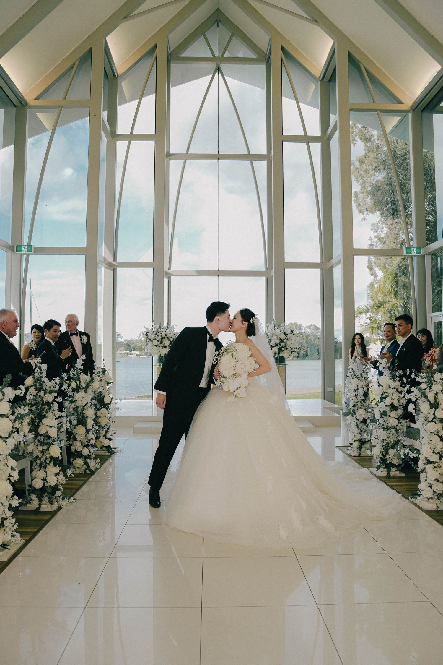 A panoramic view of a modern glass chapel wedding ceremony with white floral arrangements and ocean views in the background.