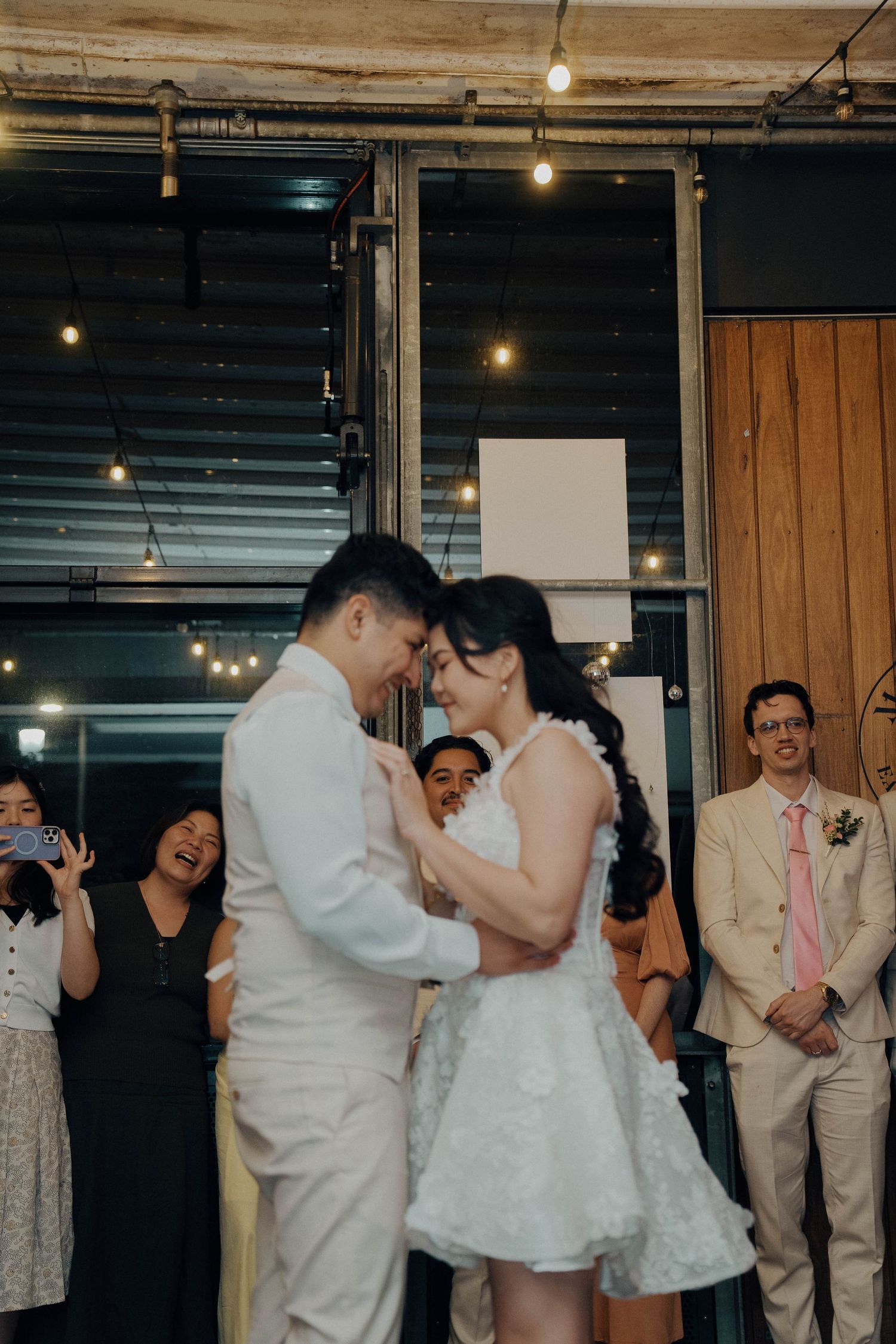 Wedding guests dance and celebrate together on the dance floor at an indoor reception venue with rustic wooden walls.