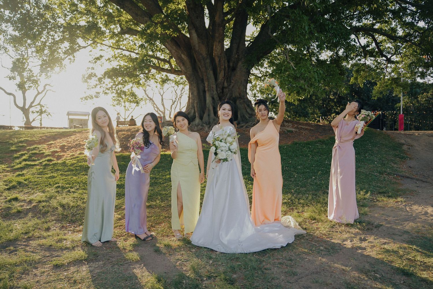 Wedding party in pastel bridesmaid dresses poses together outdoors at sunset in a garden setting.
