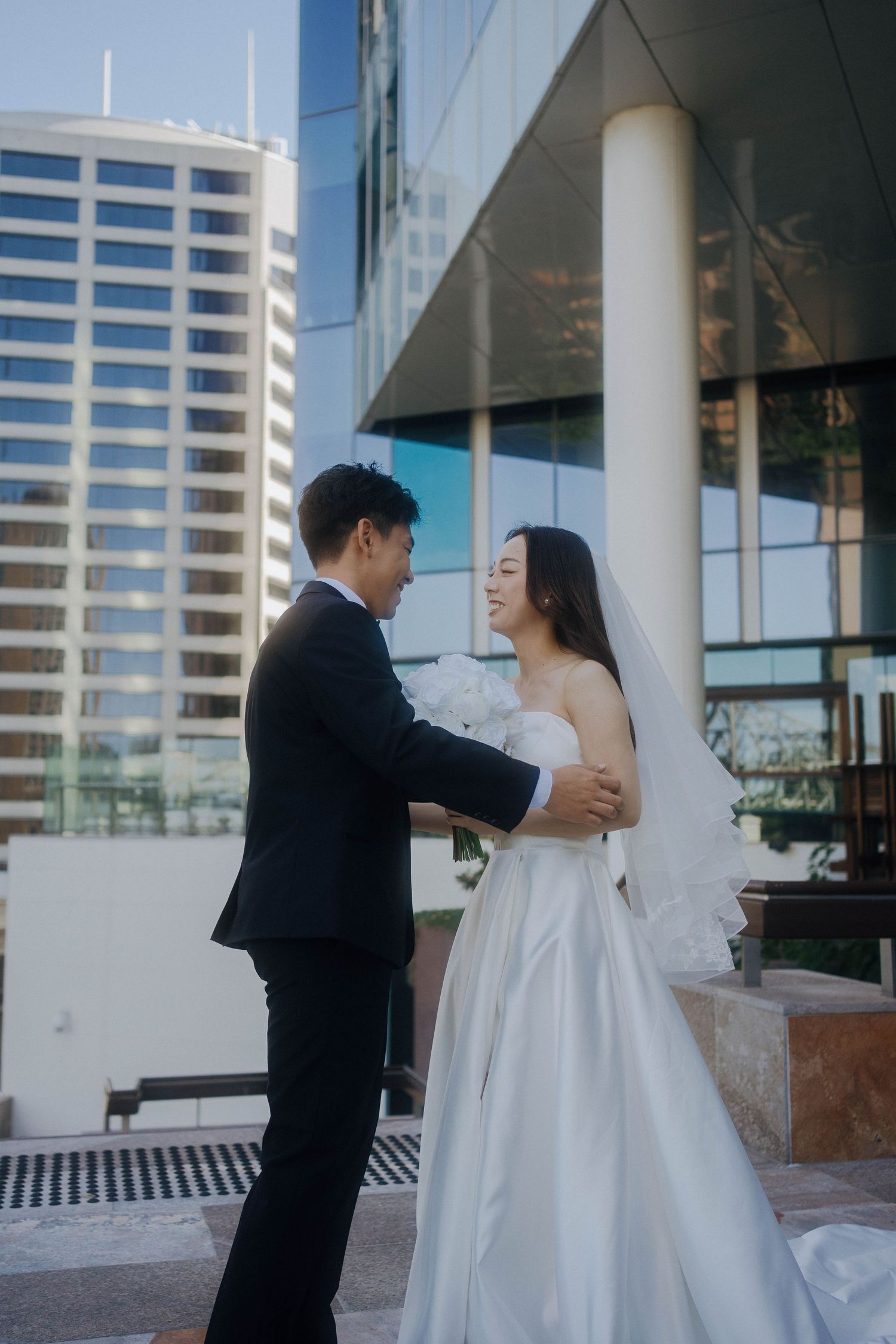 A couple in wedding attire share an intimate moment outside a modern building with glass windows and white columns.