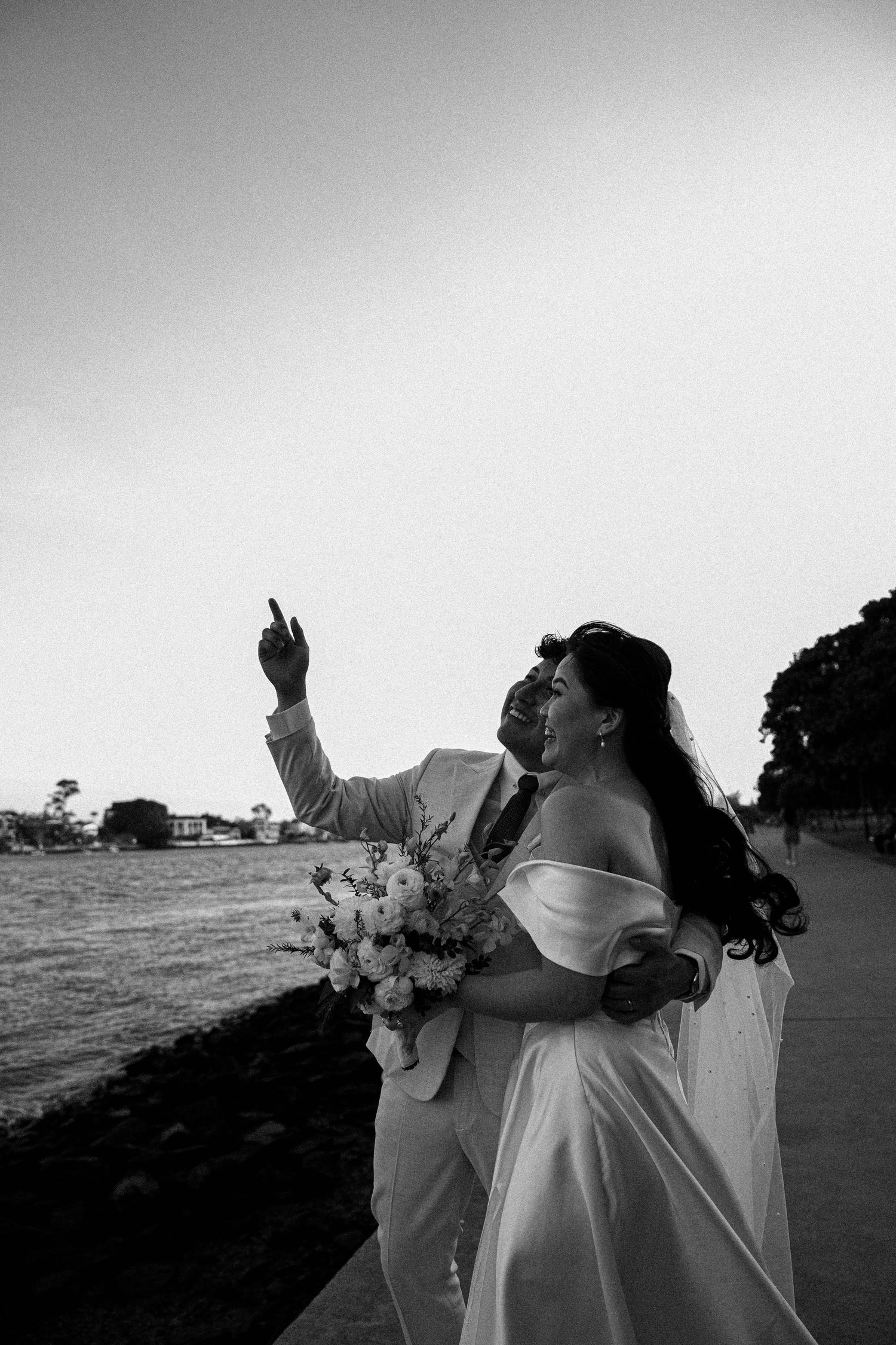 A bride in a flowing white dress holds a bouquet and gestures upward during an outdoor black and white wedding photo.
