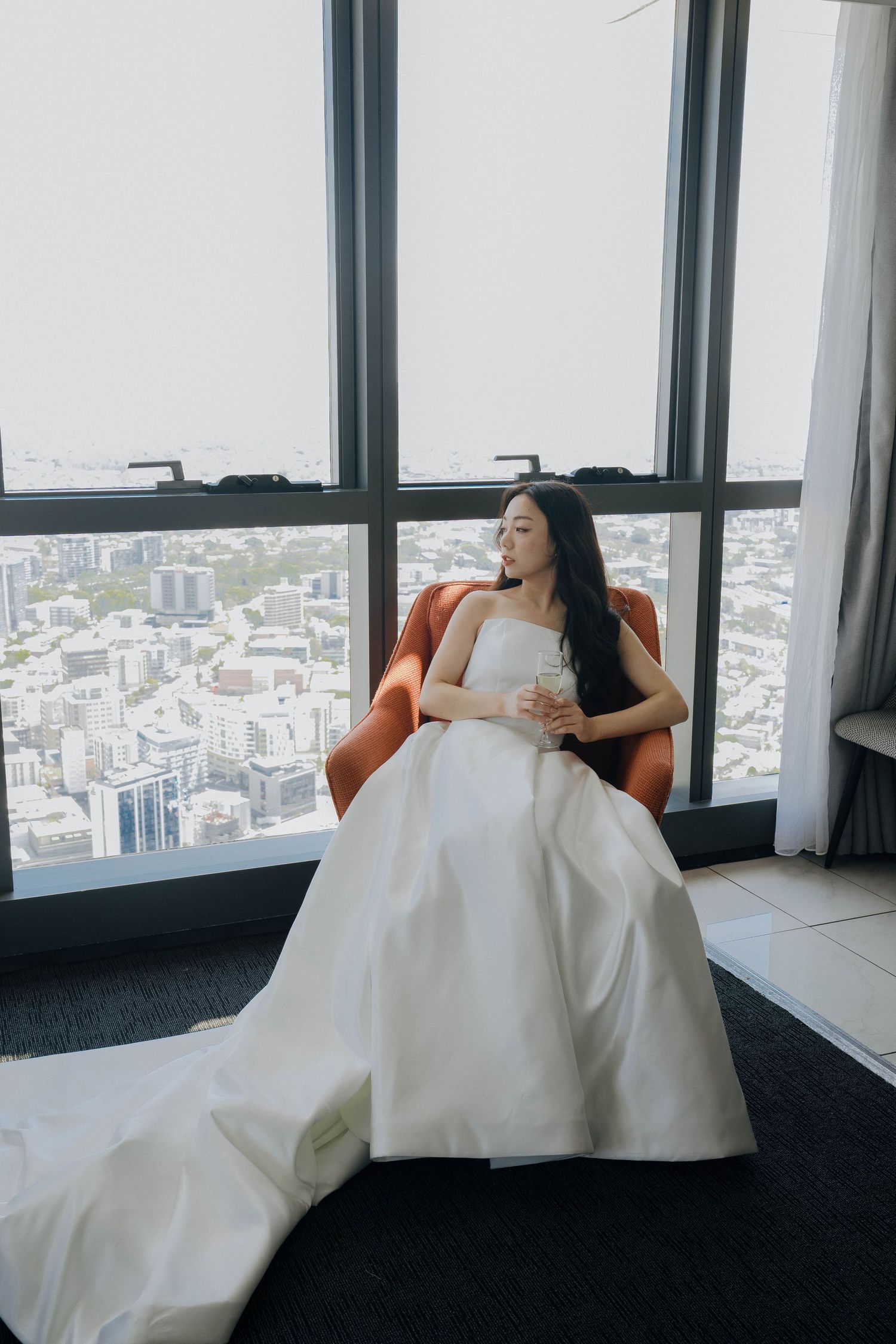 A person in an elegant white wedding gown sits in an orange chair by a high-rise window overlooking a cityscape.