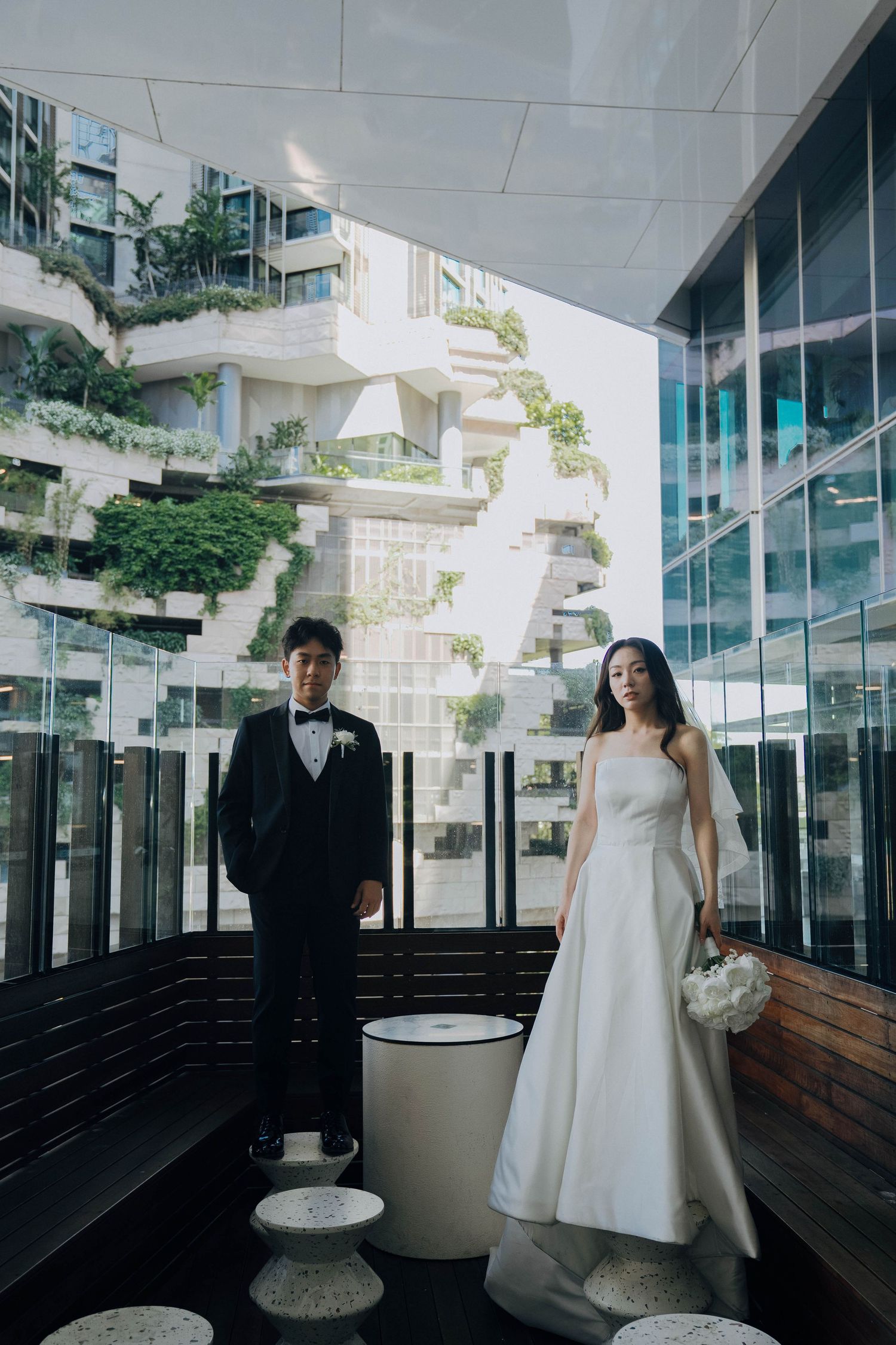Modern wedding couple poses in stylish architectural setting with glass walls and cascading greenery.