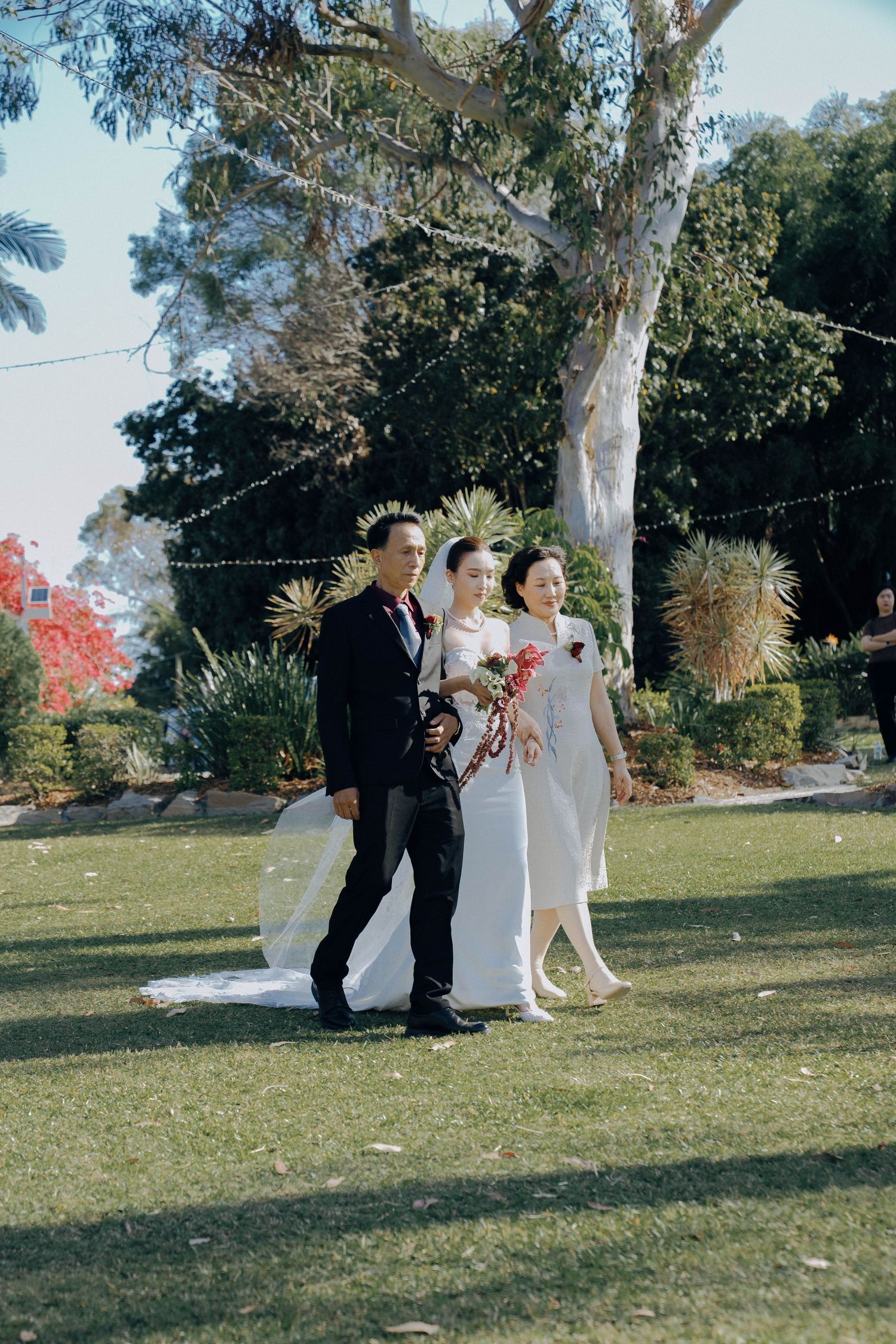 Wedding party in formal attire walks across a sunny lawn with autumn trees in the background.