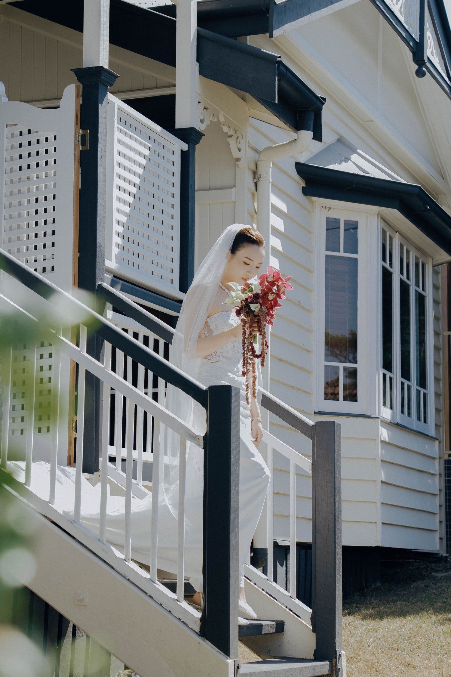 A bride in a white dress and veil stands on a staircase of a white house holding a cascading bouquet.