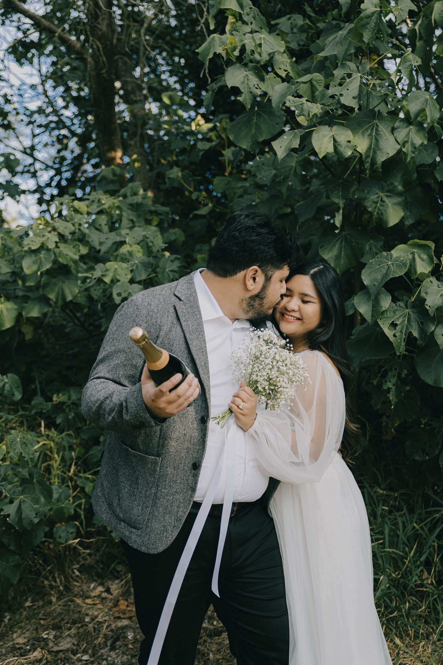 A couple shares an intimate moment outdoors among green foliage while holding a small bouquet of baby's breath flowers.