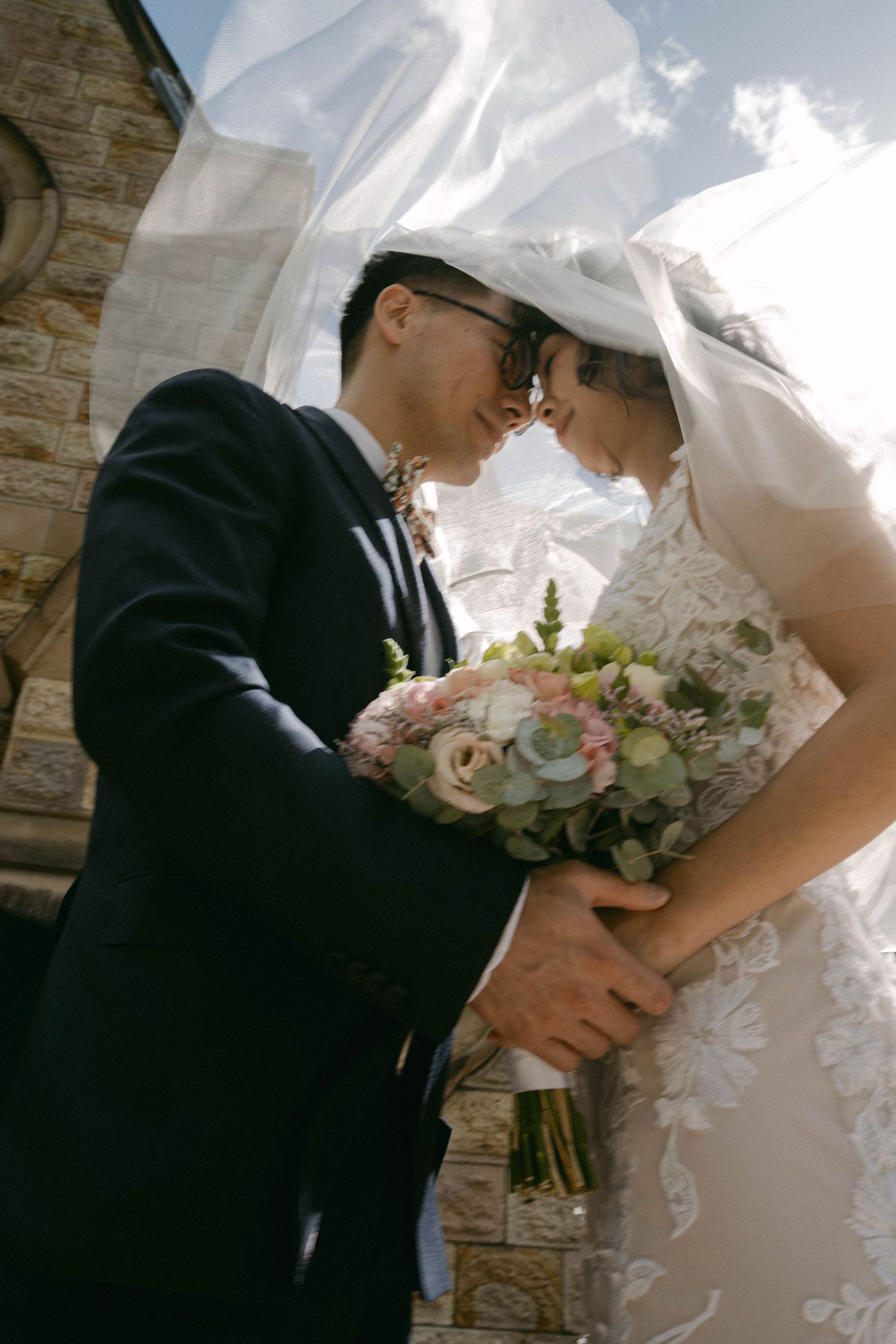 Bride and groom share an intimate moment under bridal veil while holding a soft pink and white floral bouquet.