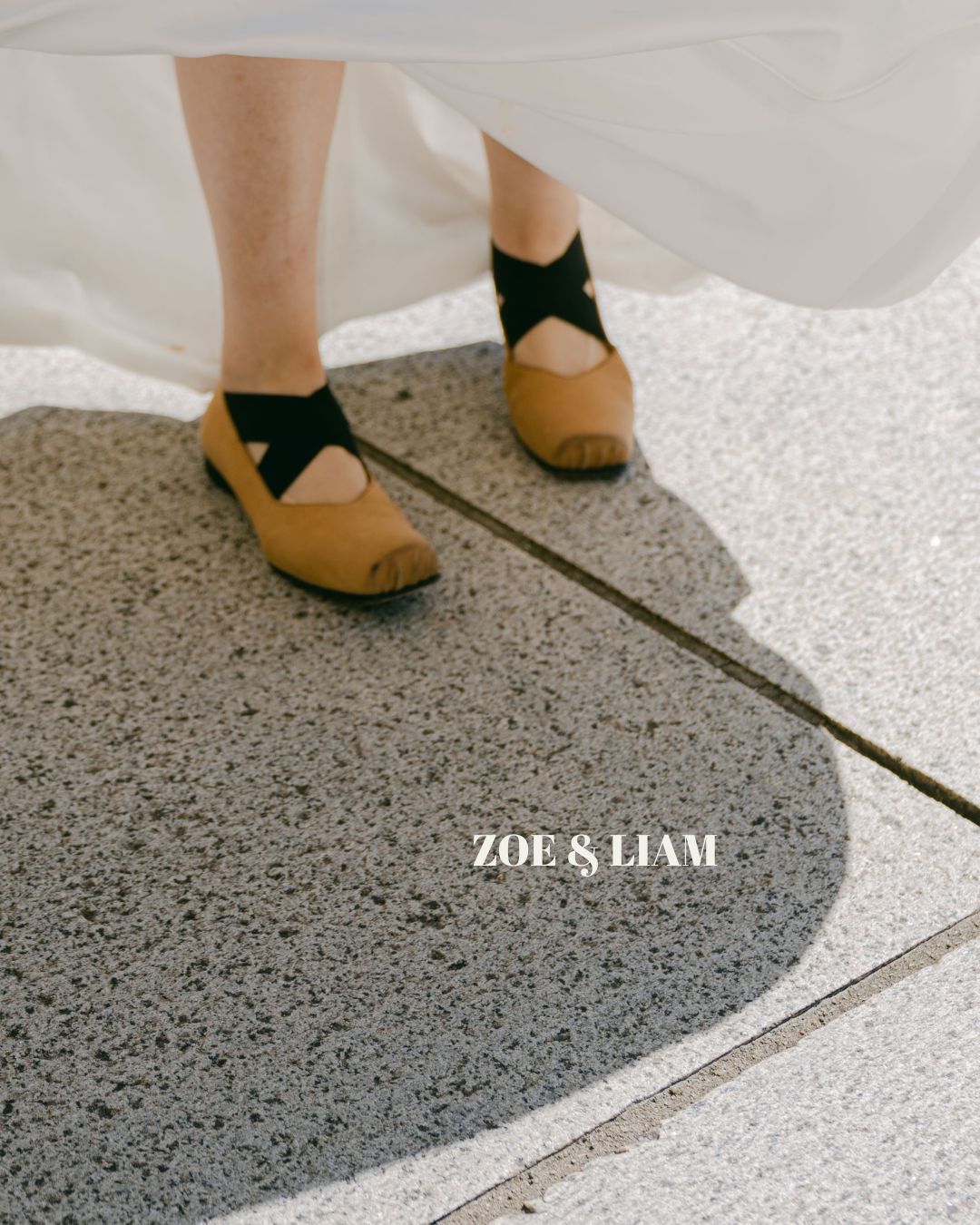 A pair of tan leather ballet flats with black bow straps photographed on a grey speckled floor tile with white edging.