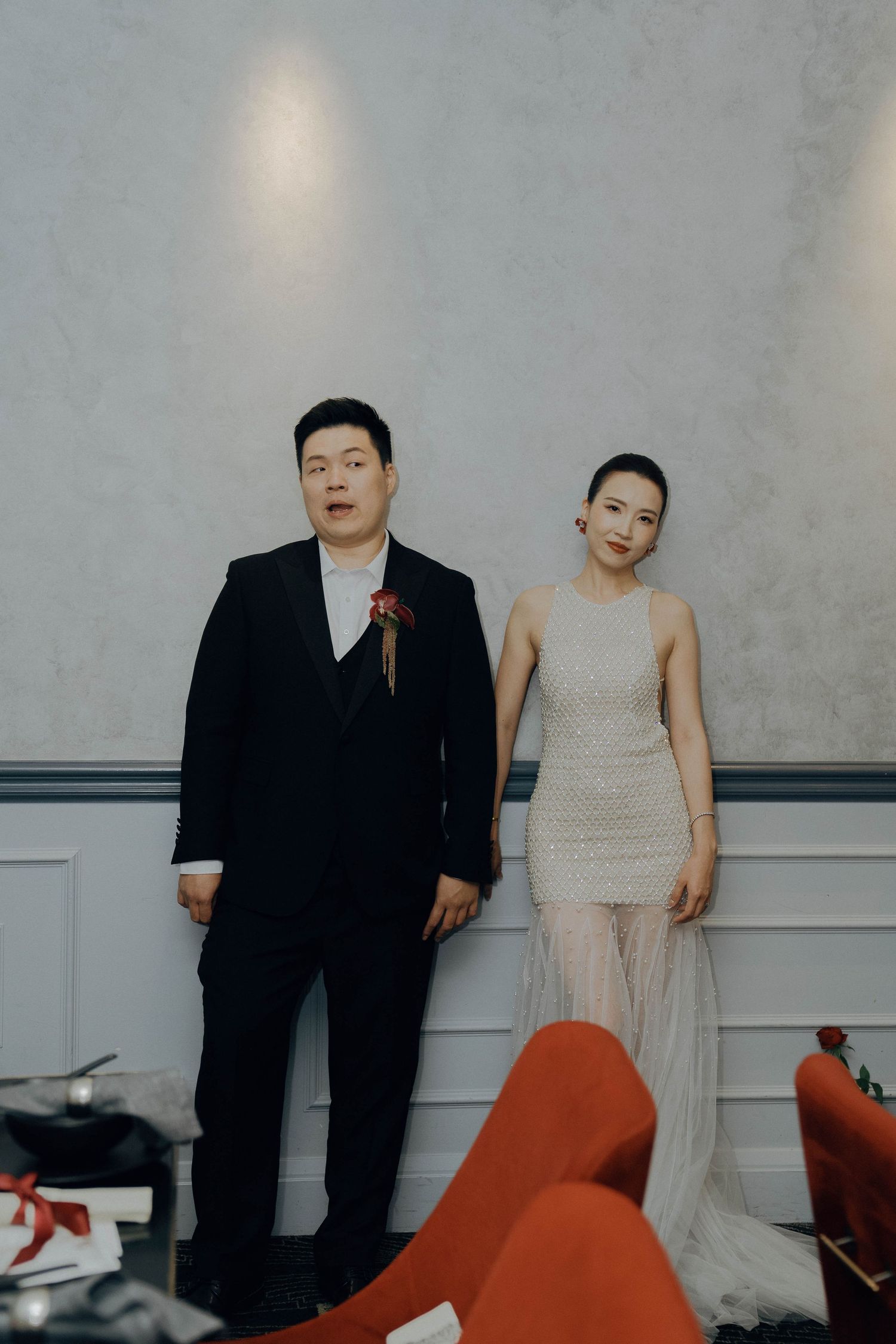 Two people pose together against a gray wall with red furniture visible in the foreground.