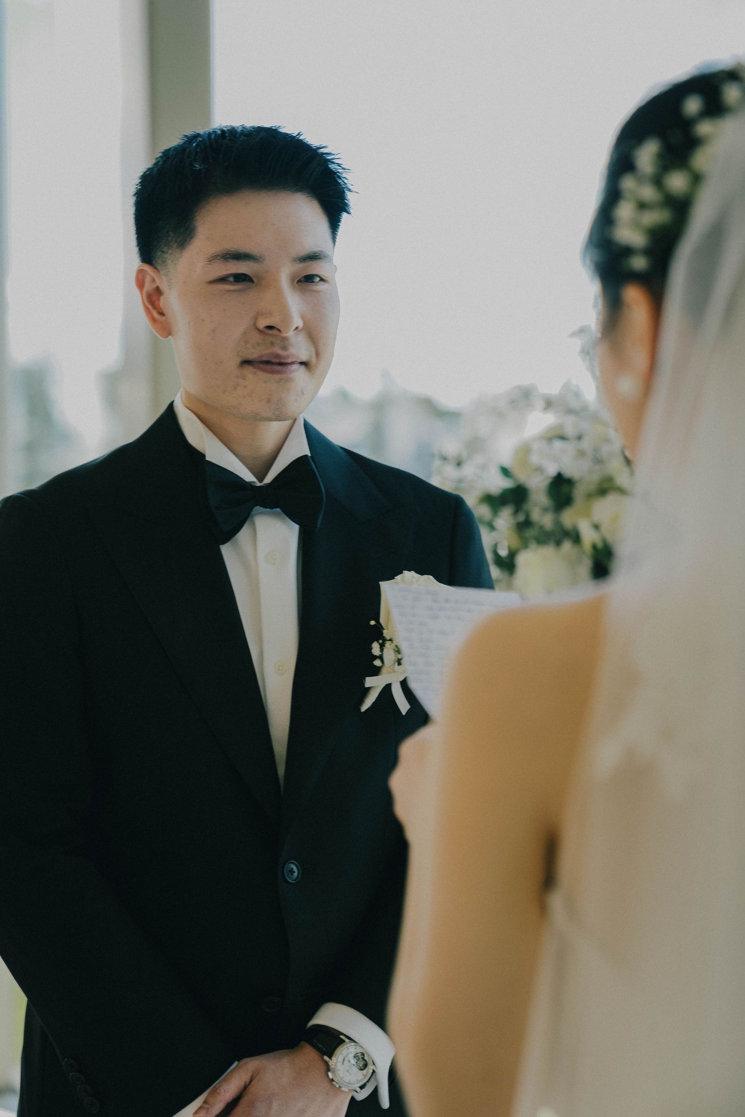 A groom in a black tuxedo and bowtie smiles during a wedding ceremony with city views in the background.