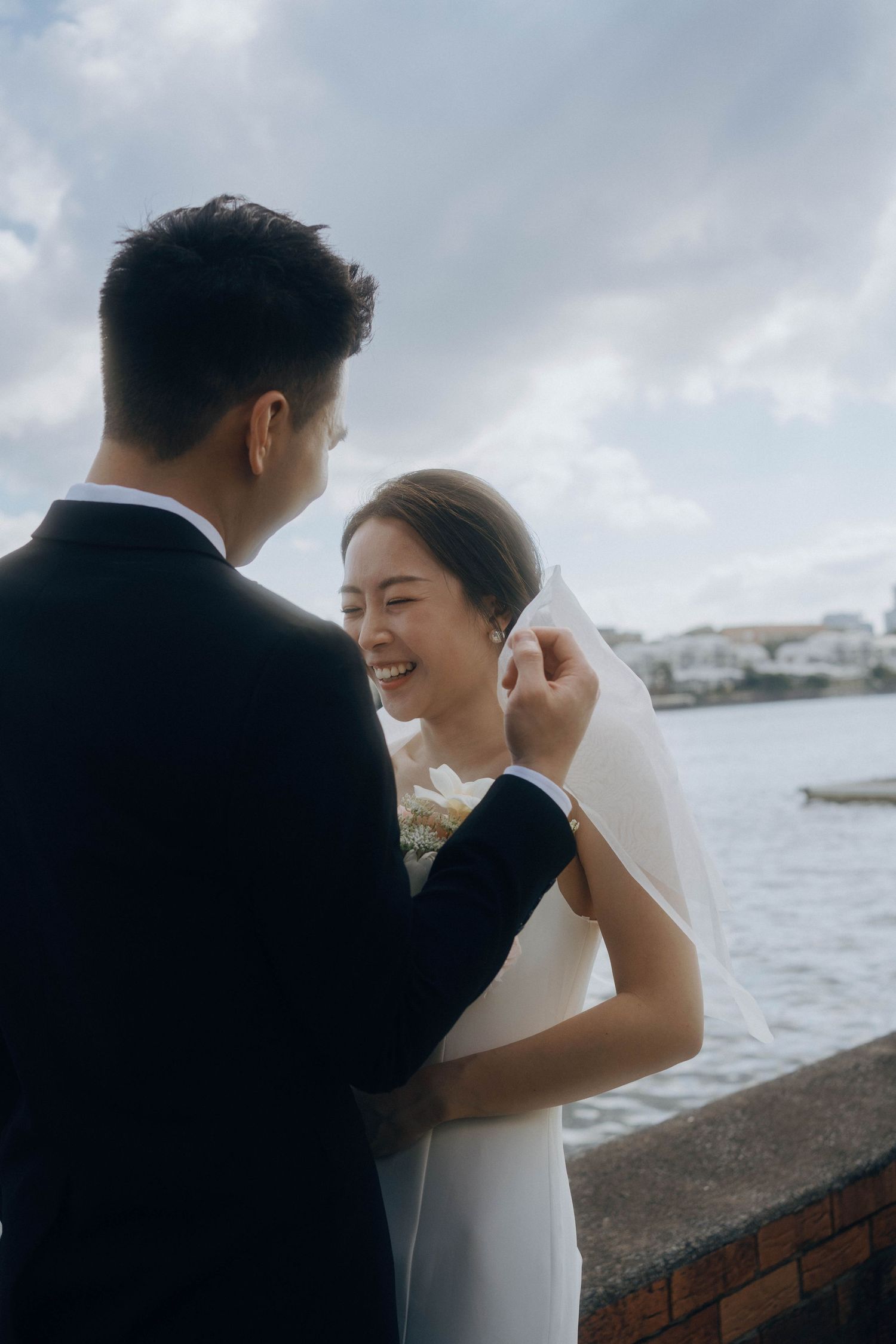 A romantic wedding couple embraces by the waterfront on a cloudy day, with white buildings visible in the background.