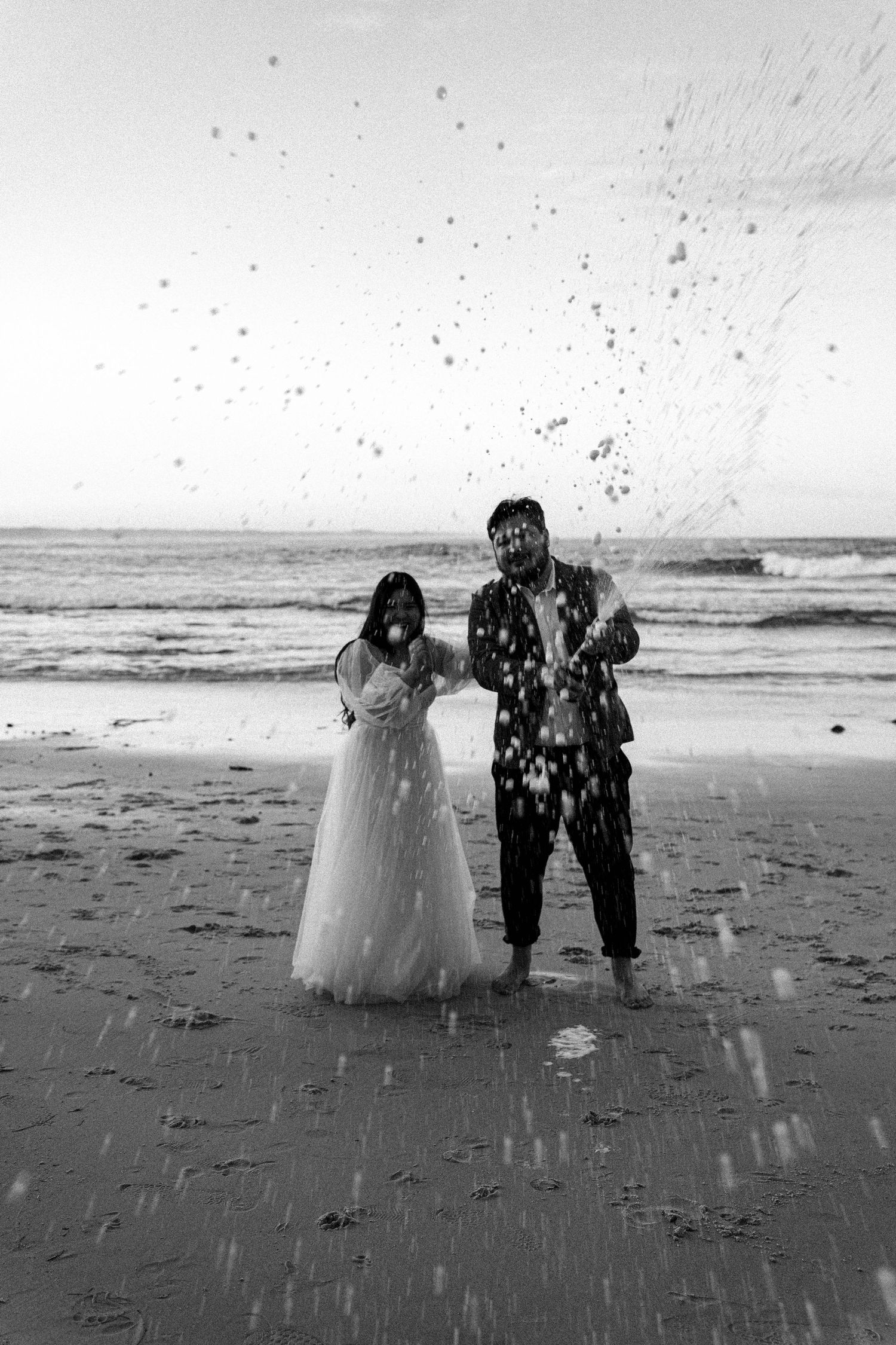 A couple in wedding attire playfully splashes water on a beach in black and white photography.
