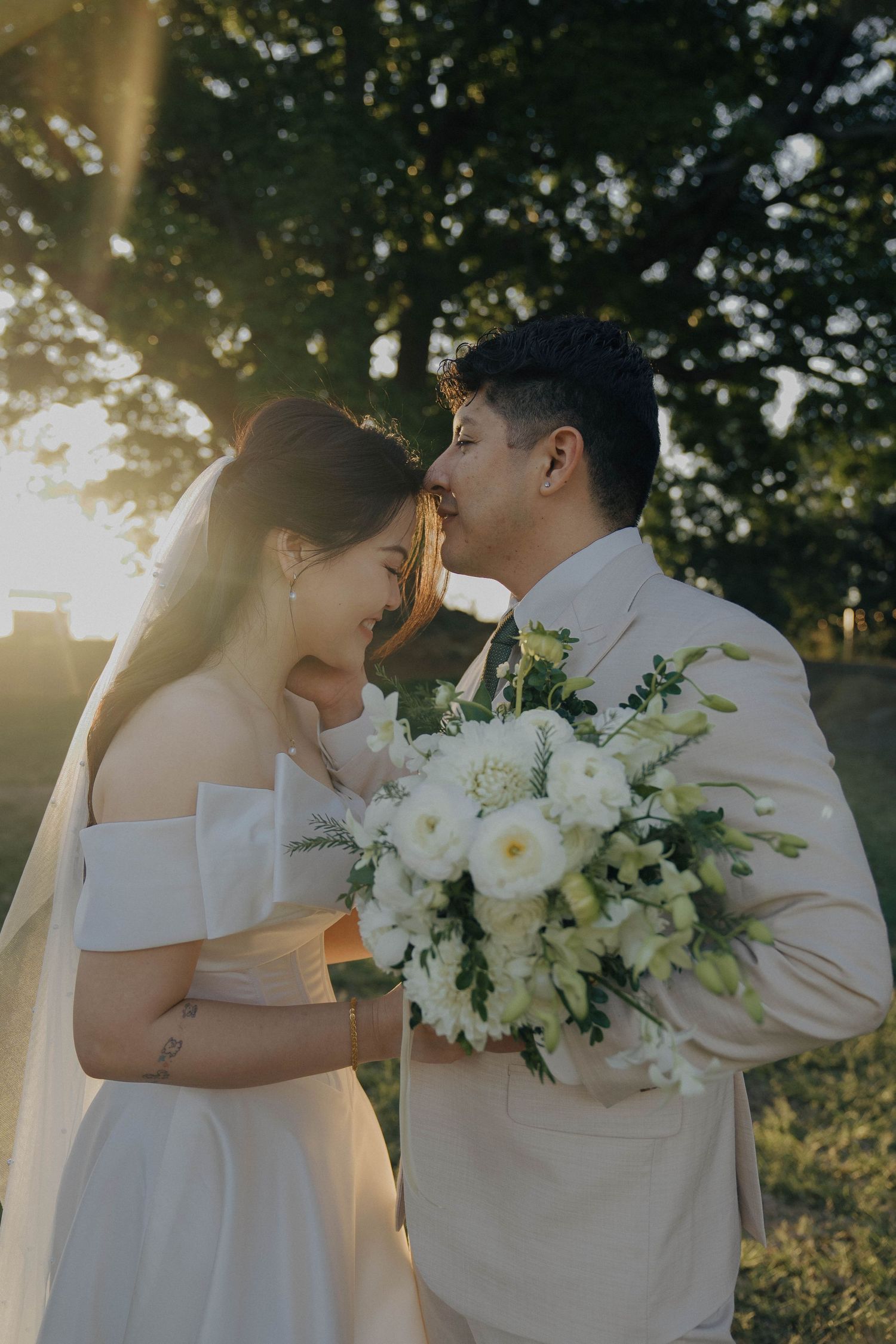 Romantic wedding couple embraces in golden sunlight with white floral bouquet in an outdoor garden setting.