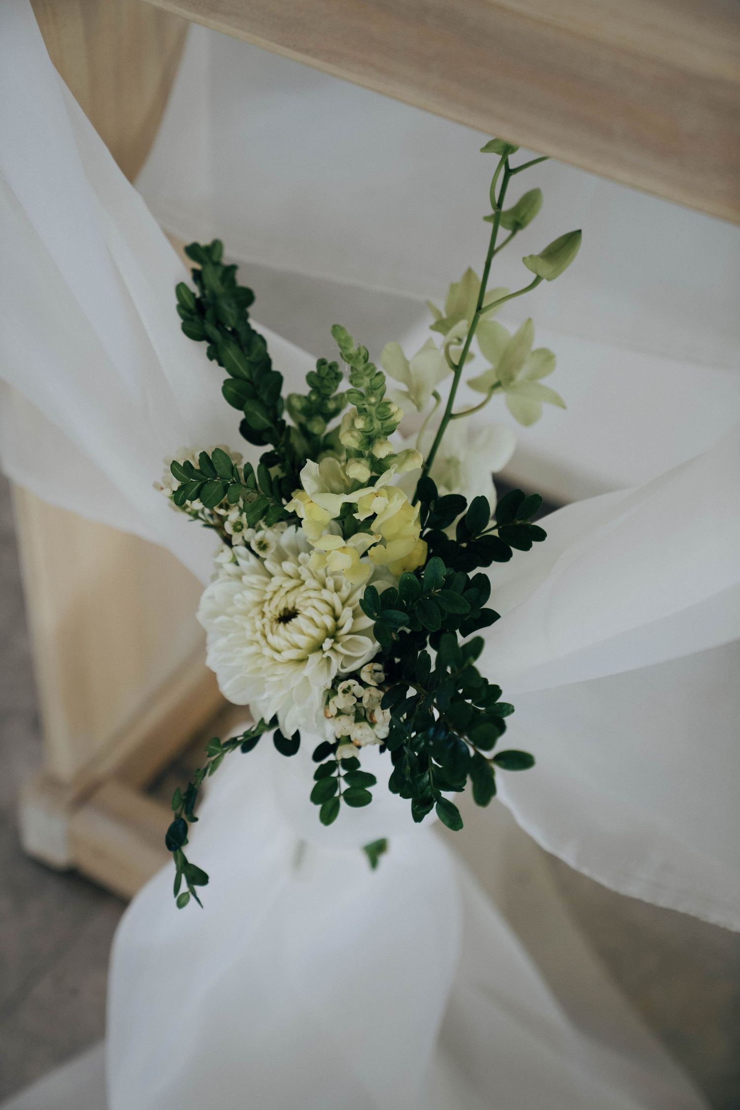 Elegant white floral arrangement with green foliage against draped white fabric creating a soft, romantic wedding decor detail.