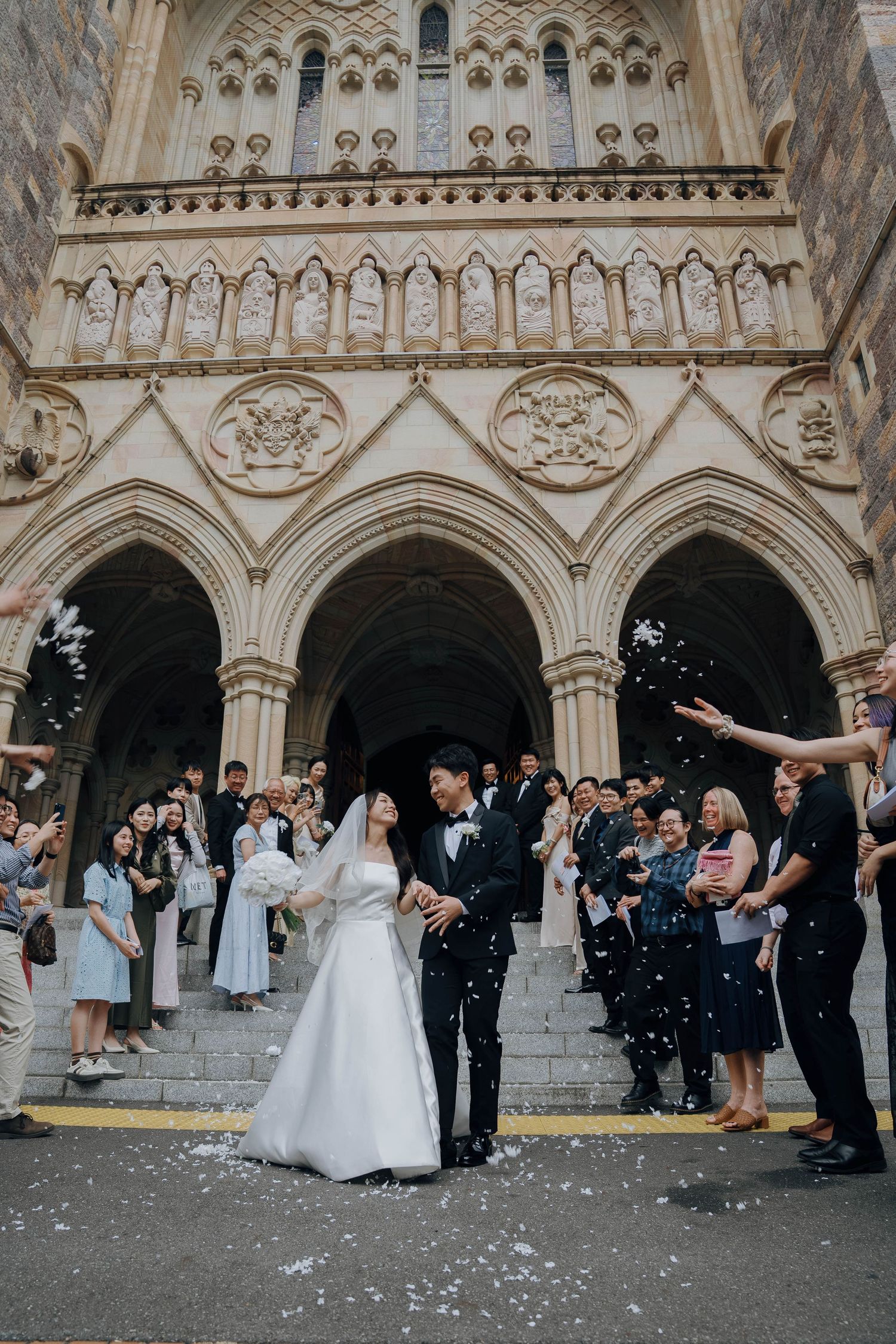 Wedding celebration outside a Gothic cathedral with ornate stone arches as guests throw confetti on the church steps.