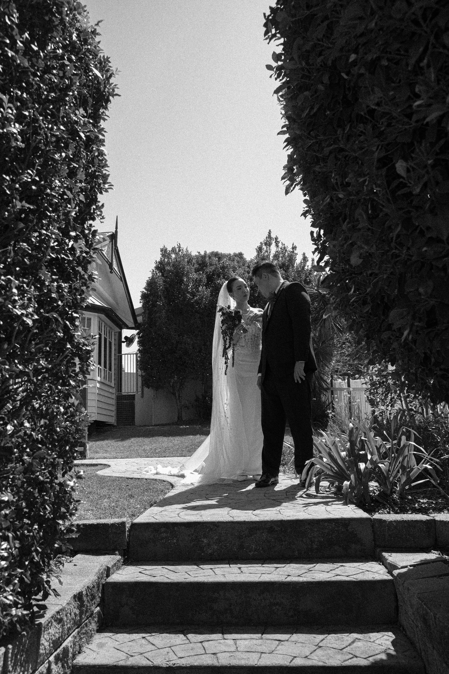 A couple in wedding attire walks along a stone path surrounded by lush gardens in black and white photography.